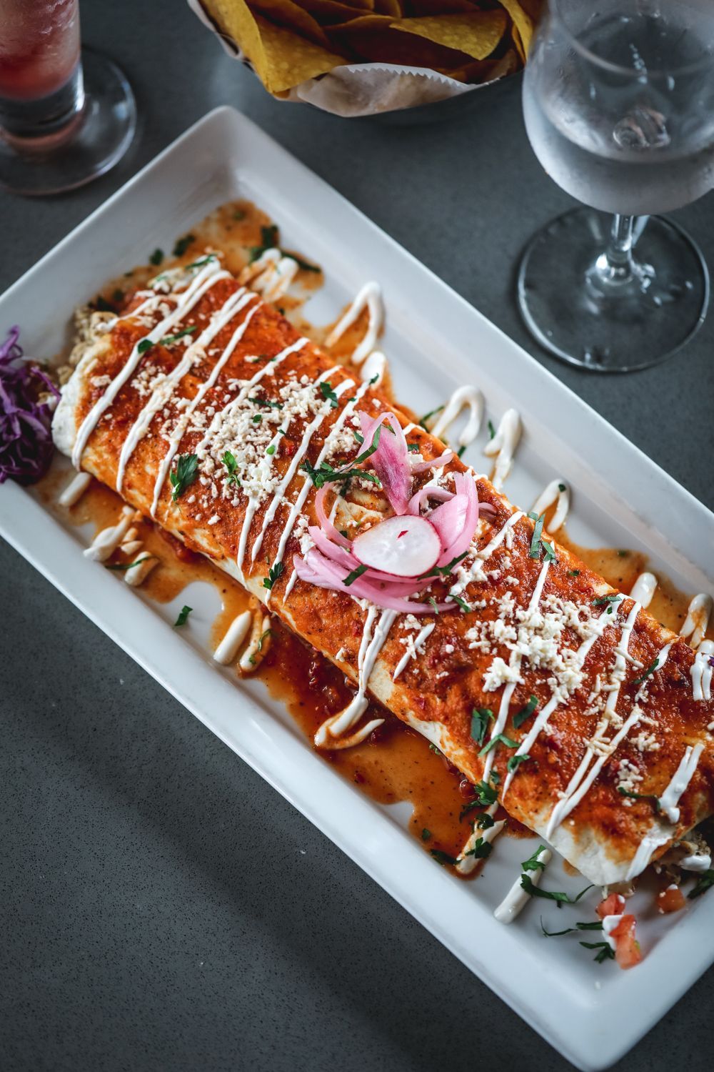 Enchilada-style burrito on a rectangular white plate, drizzled with white sauce, garnished with pickled onions and crumbled cheese.