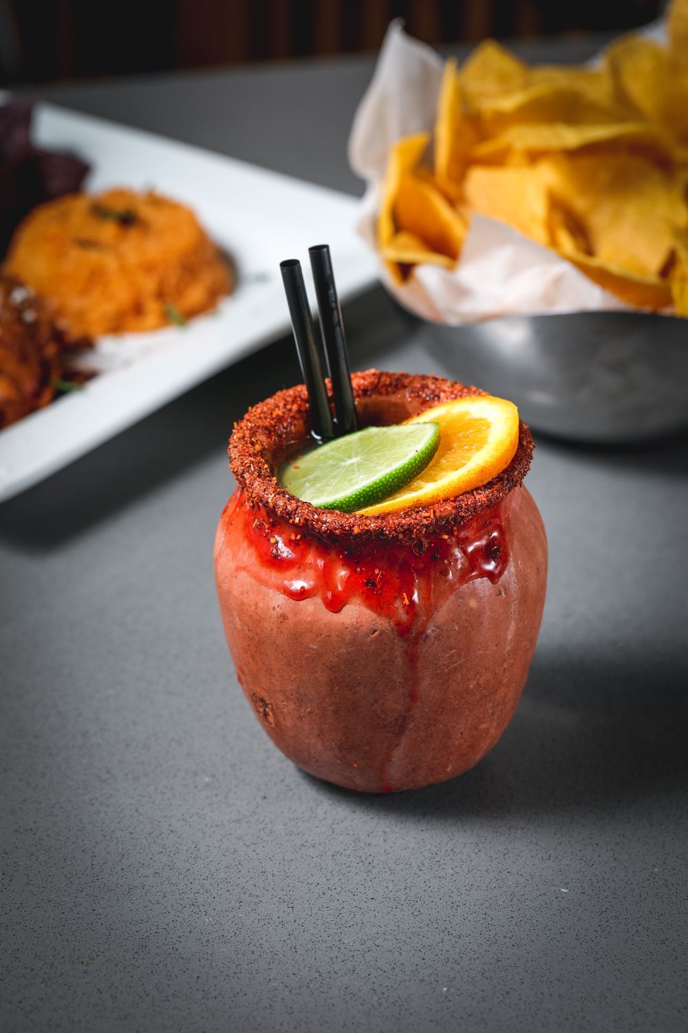 Cocktail in a clay cup, rimmed with chili, lime and orange garnish, two straws, with chips and food in background.