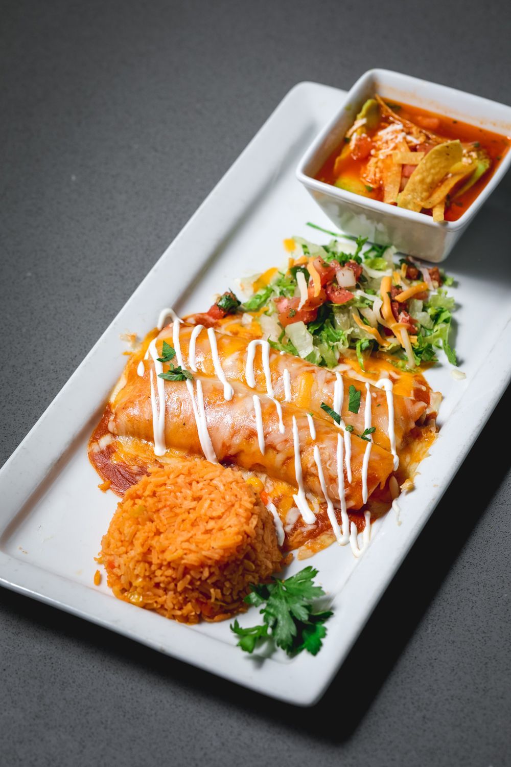 Enchilada with red sauce, rice, salad, and avocado soup on a white rectangular plate.