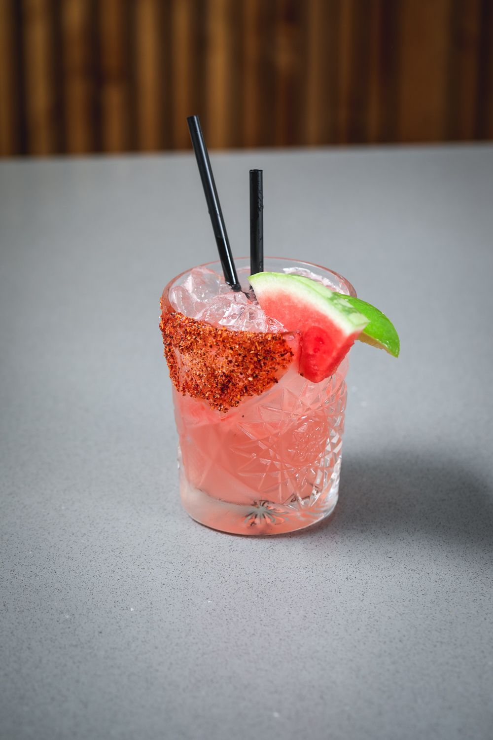 Watermelon cocktail with chili rim, ice, and black straws. Garnished with a watermelon slice on a grey surface.