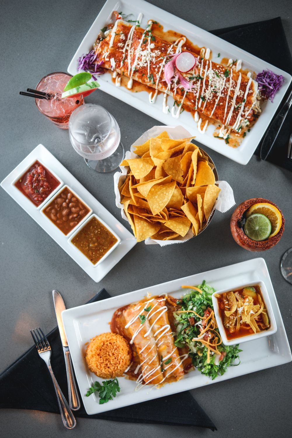 Mexican food spread: enchiladas, salsa, chips, and a dish with rice and garnish. Drinks are included.