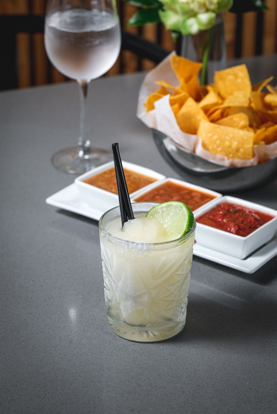 Frozen margarita with lime, salsa, chips, and water glass on a gray table.