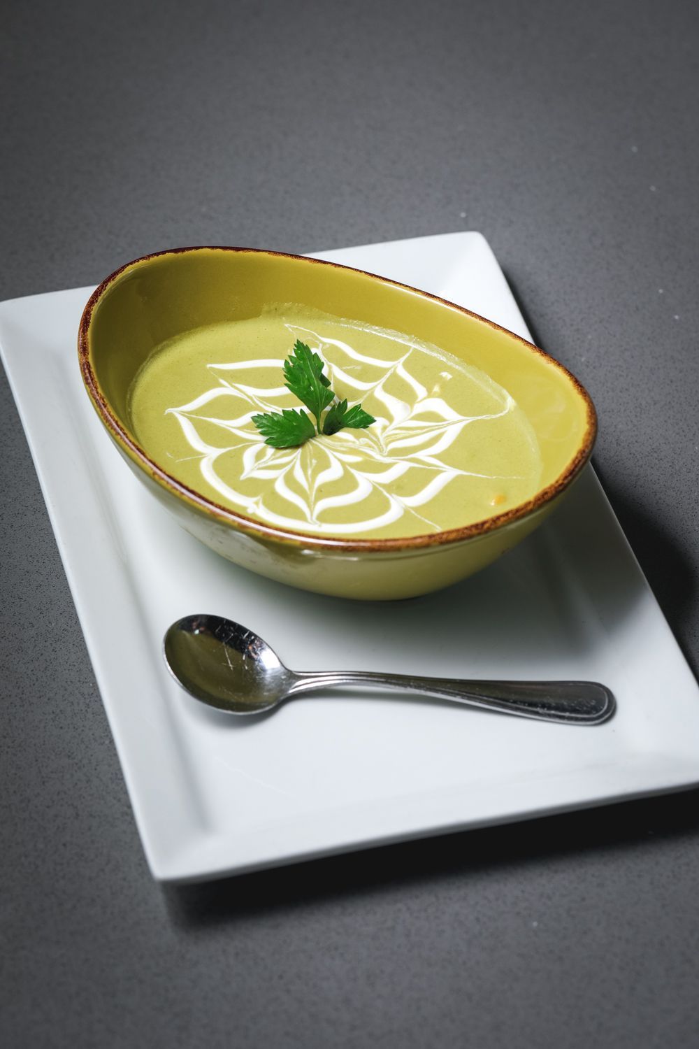 Green soup in yellow bowl on white plate with a spoon; decorated with cream swirls and parsley.