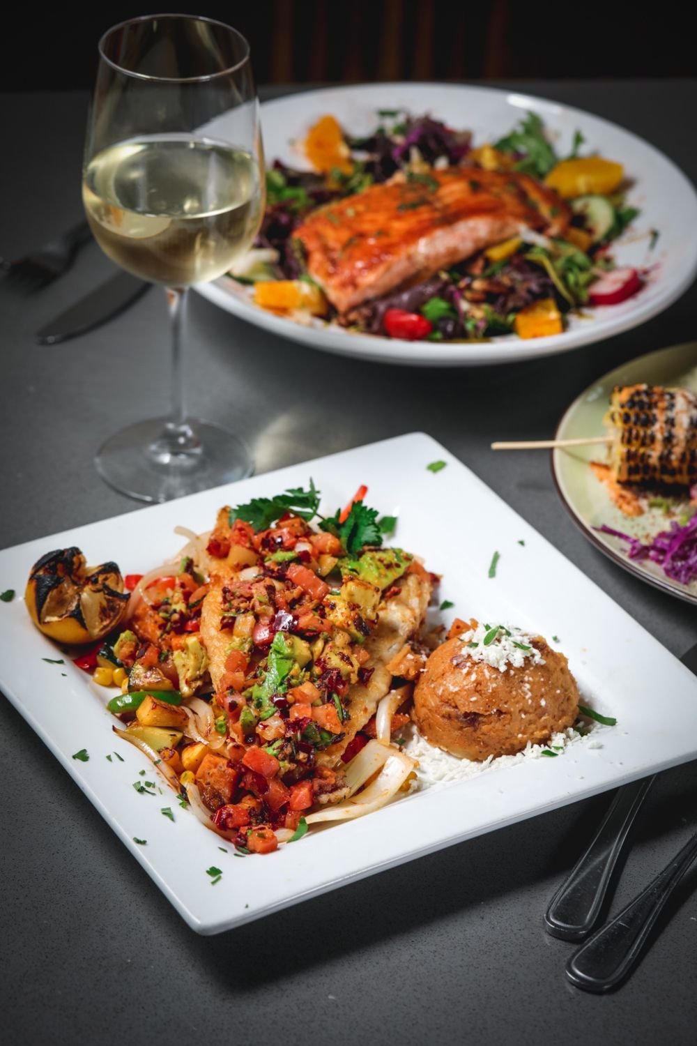 Plates of food on a table, including salmon salad, fish with salsa, corn, and a glass of white wine.
