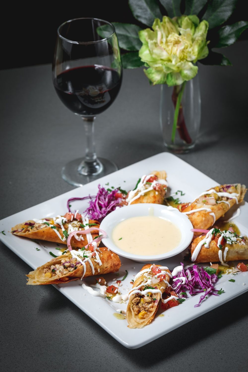 Egg rolls with dipping sauce, red wine, and a flower arrangement on a table.