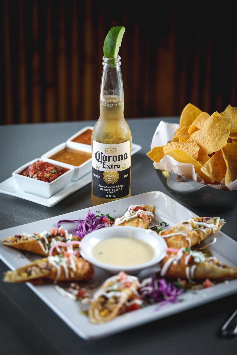 Corona beer bottle with lime, chips, salsa, fried rolls, and queso on a table.