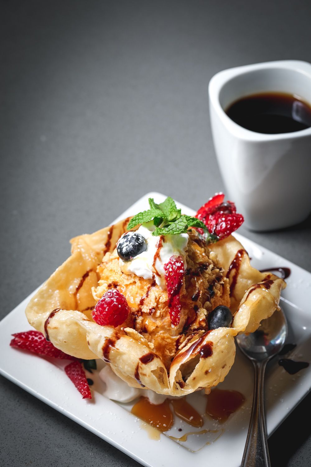 Ice cream dessert in a fried bowl, topped with berries and sauce, next to coffee.