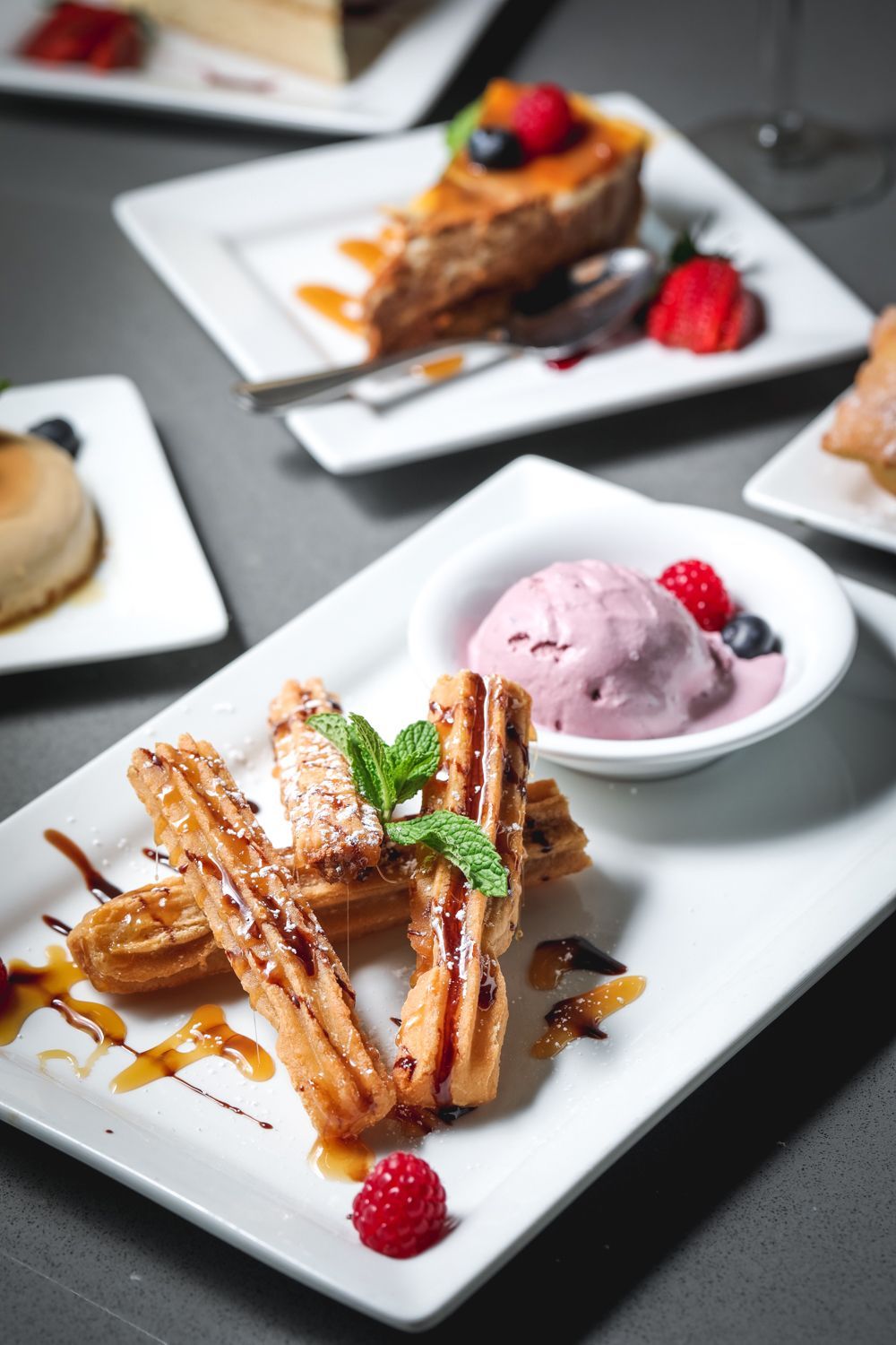 Churros drizzled with syrup and powdered sugar, served with ice cream and other desserts on white plates.