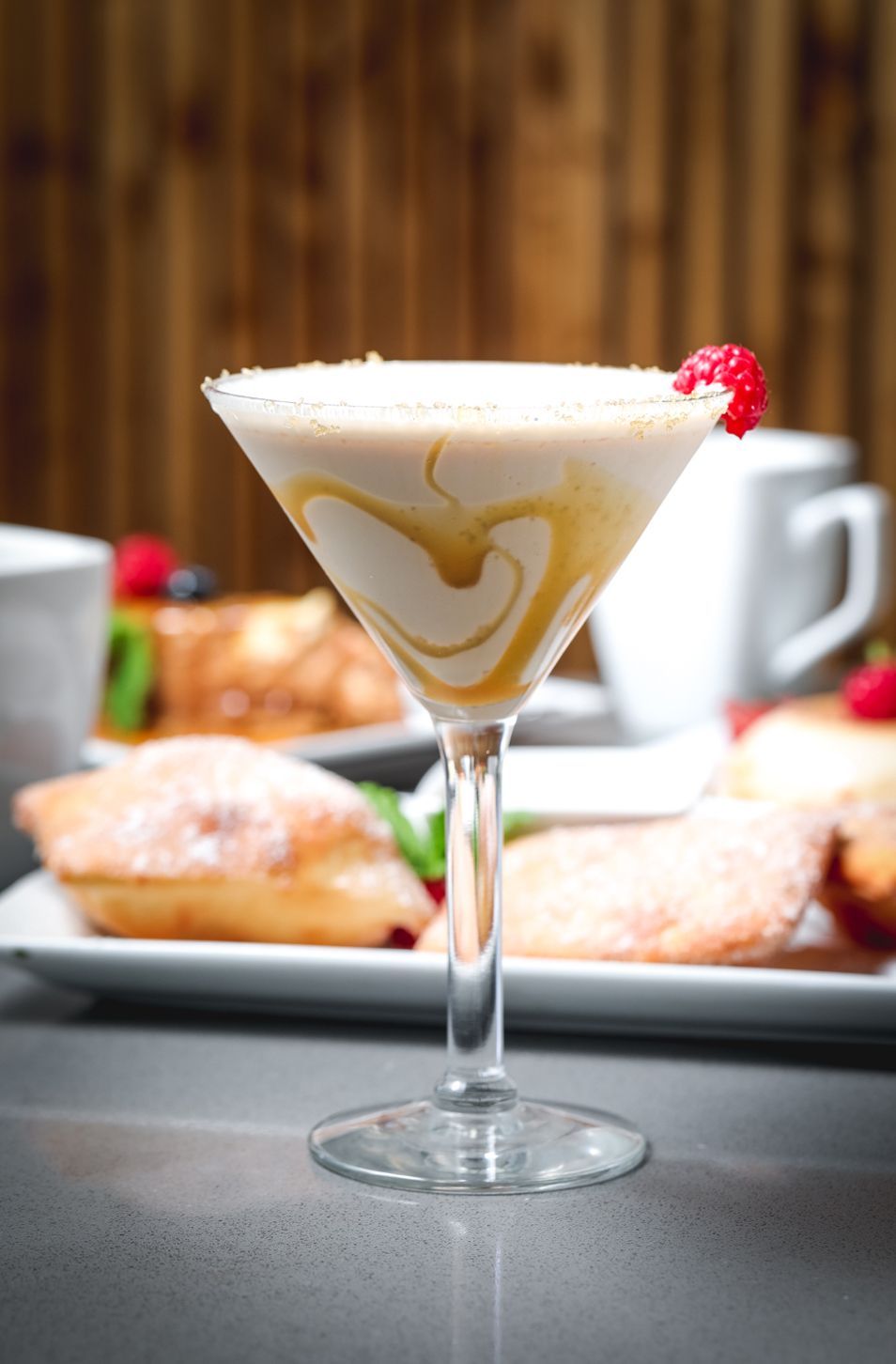 Cocktail in martini glass, swirled with caramel, garnished with raspberry. Served with pastries on a table.