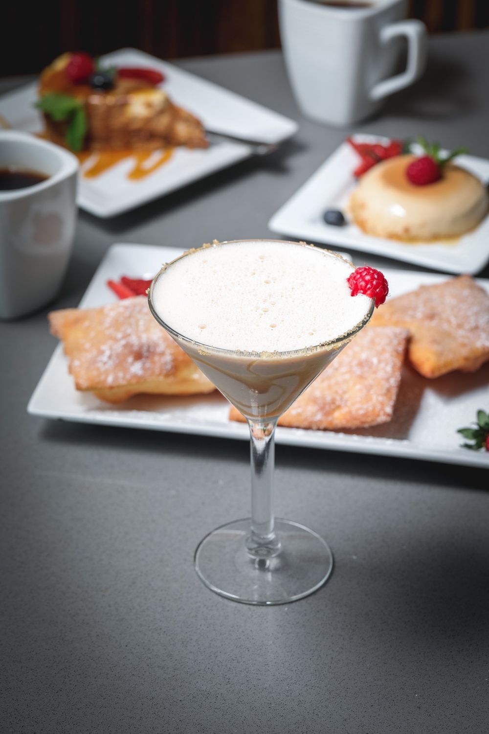 Cocktail in a martini glass with desserts on plates, coffee cups on a gray table.