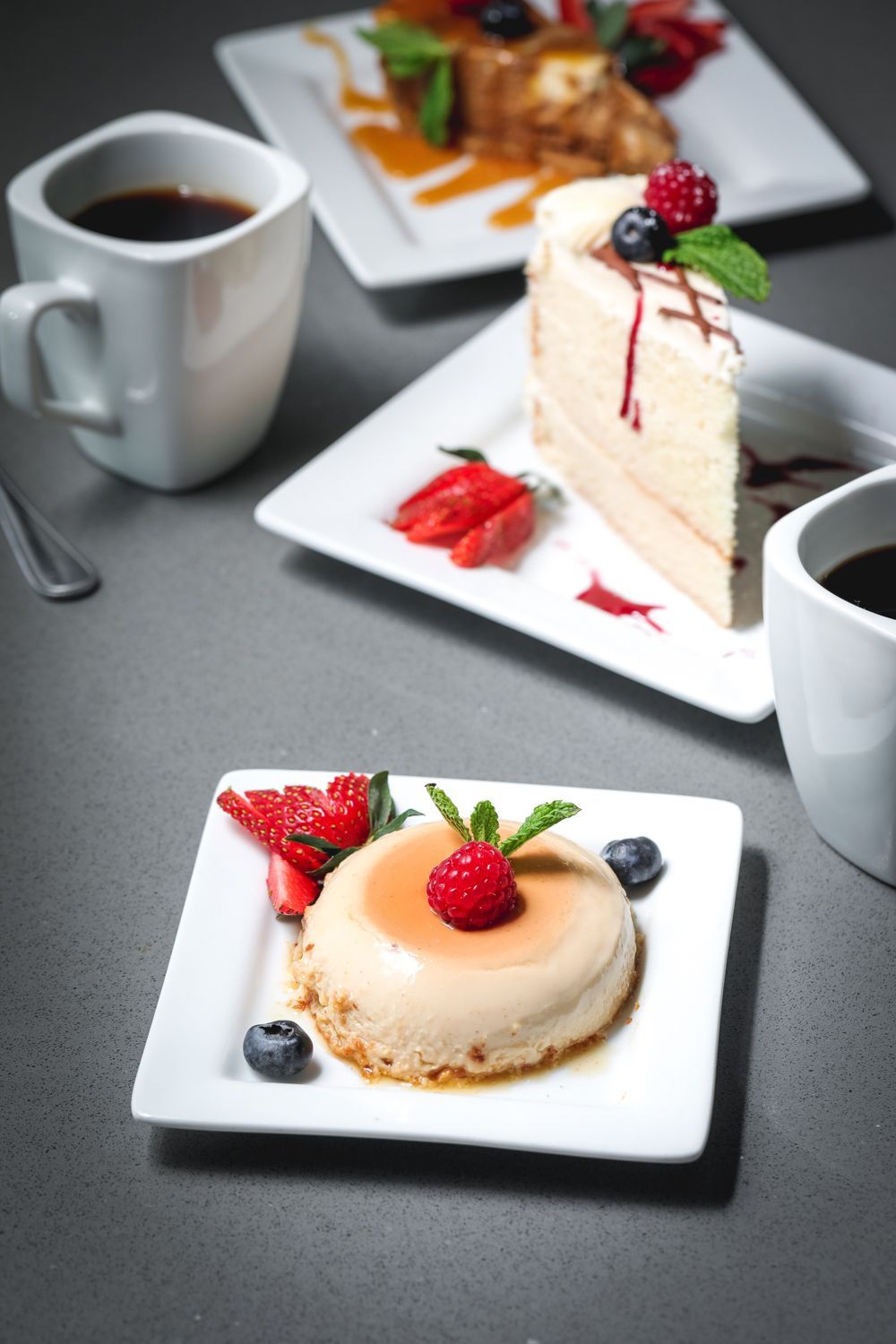 Three desserts on white plates with coffee. Includes cake, flan, and pastries, with berries and mint.