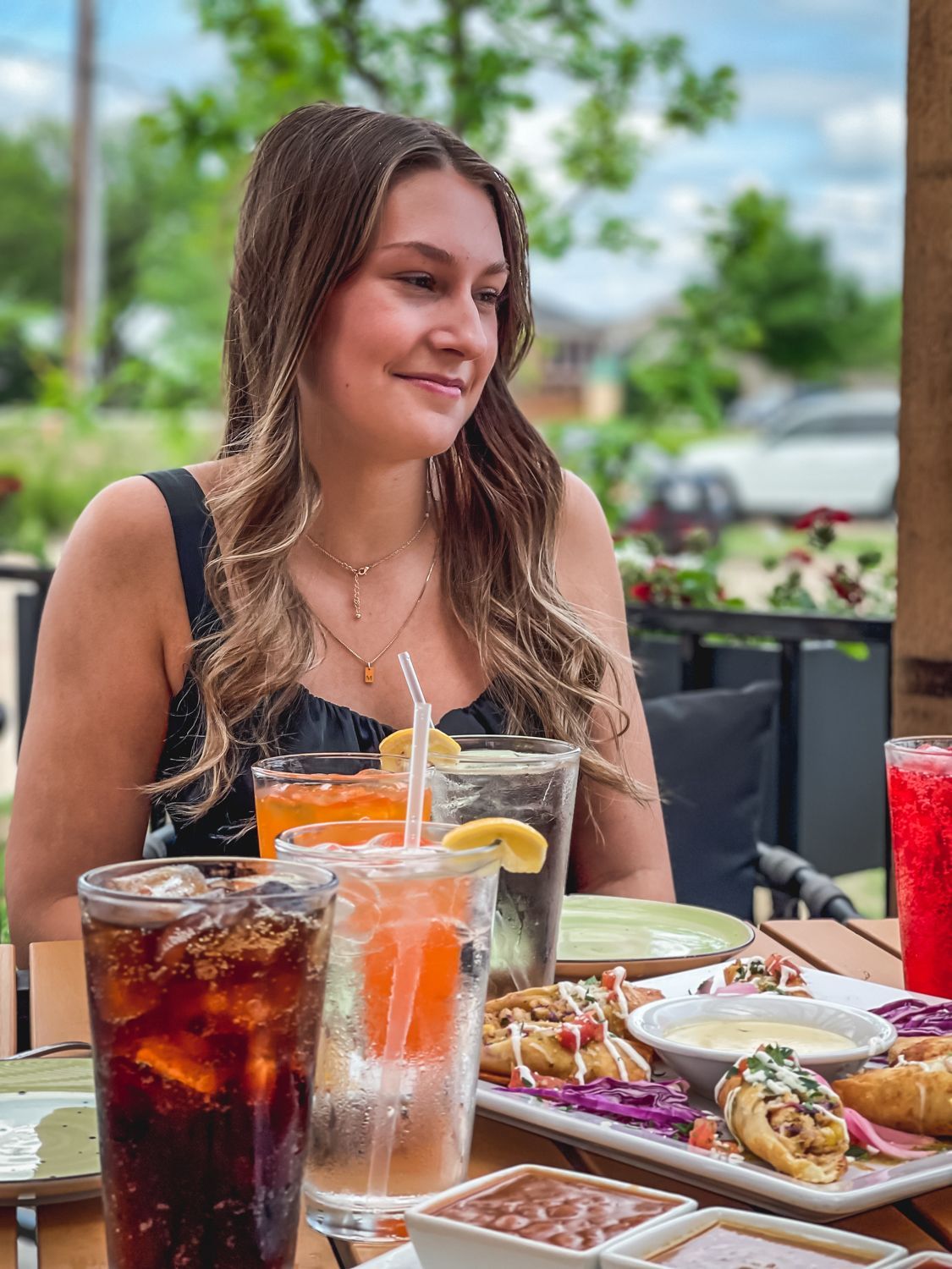 Woman at outdoor table smiles, with drinks and food.