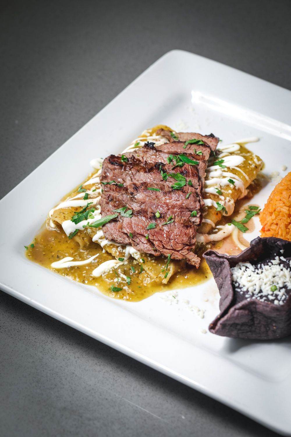 Grilled steak slices atop green enchiladas, served with rice, black beans, and cream on a white plate.