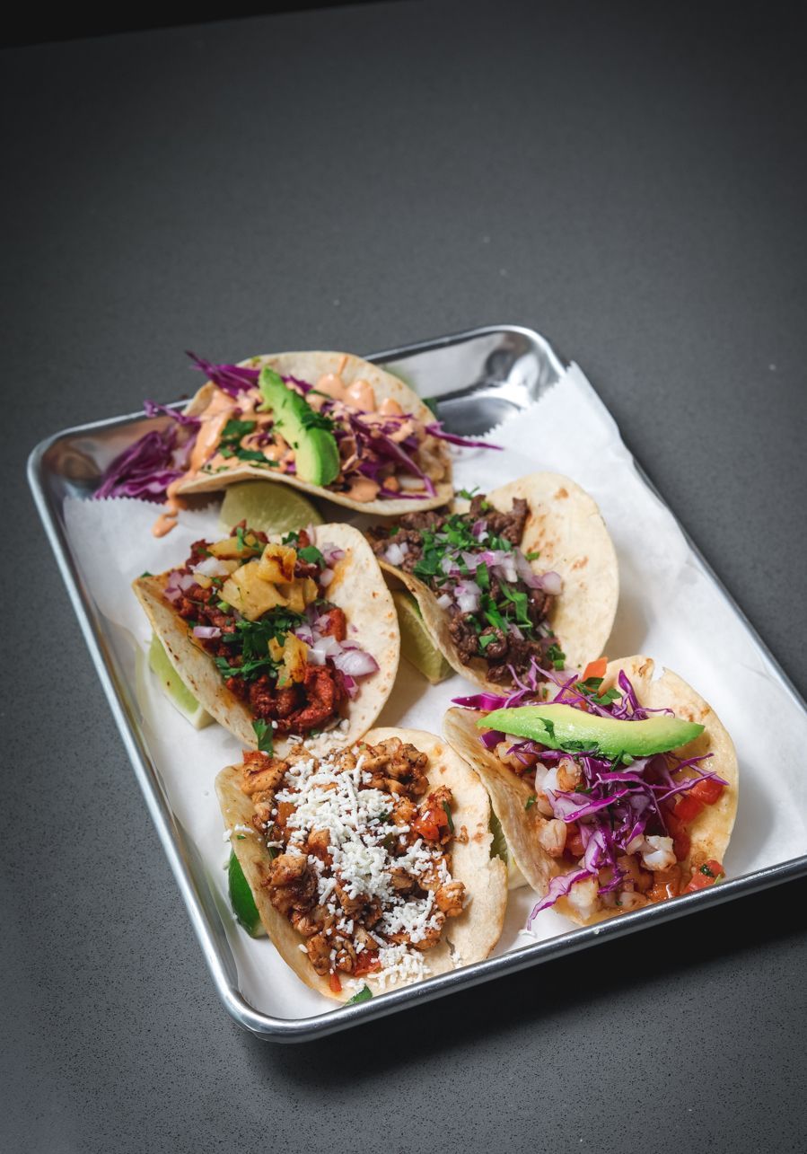 Five tacos with various toppings on a metal tray, atop a dark surface.