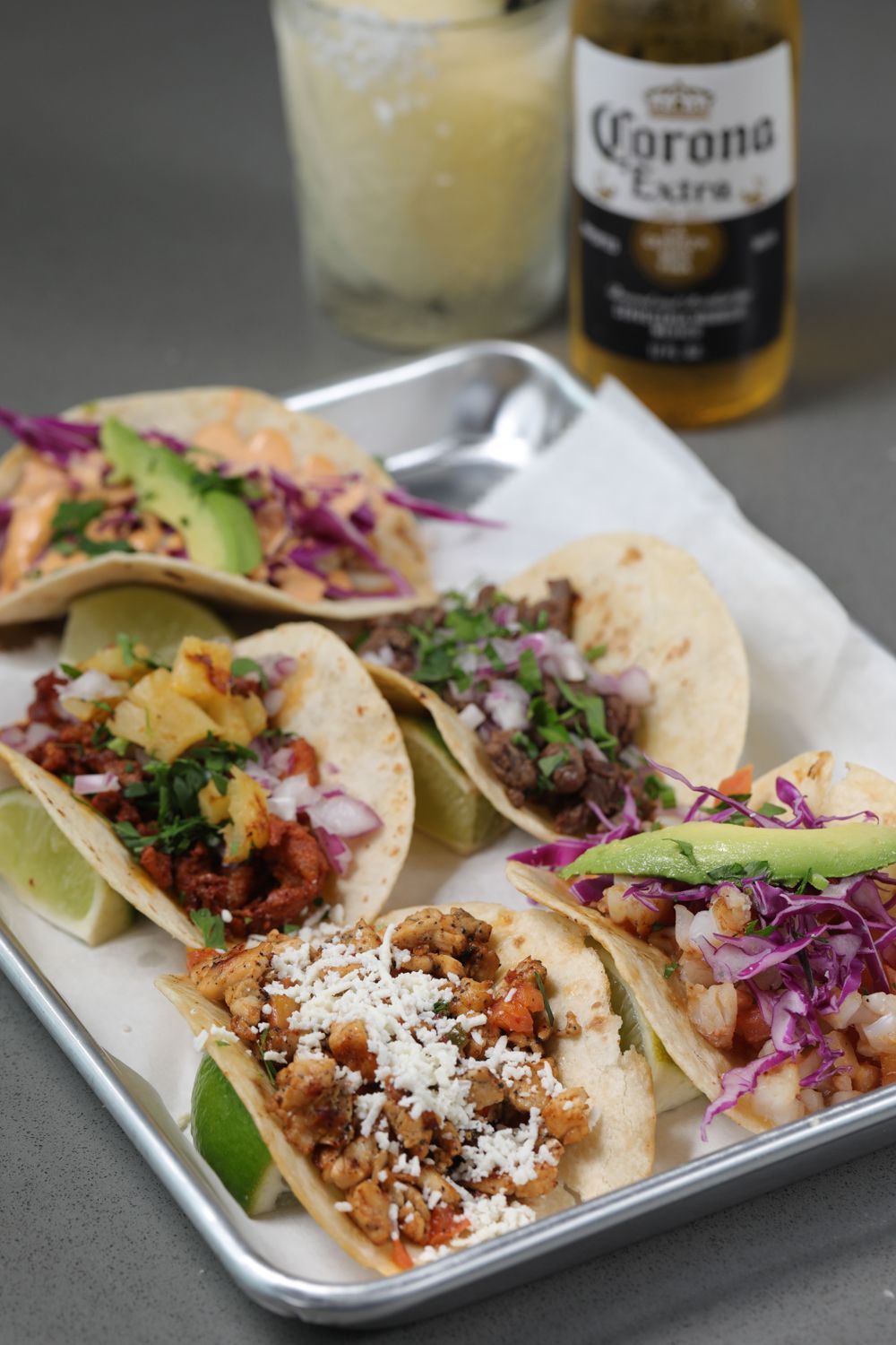 Tray of colorful tacos with a lime, margarita, and Corona beer bottle.