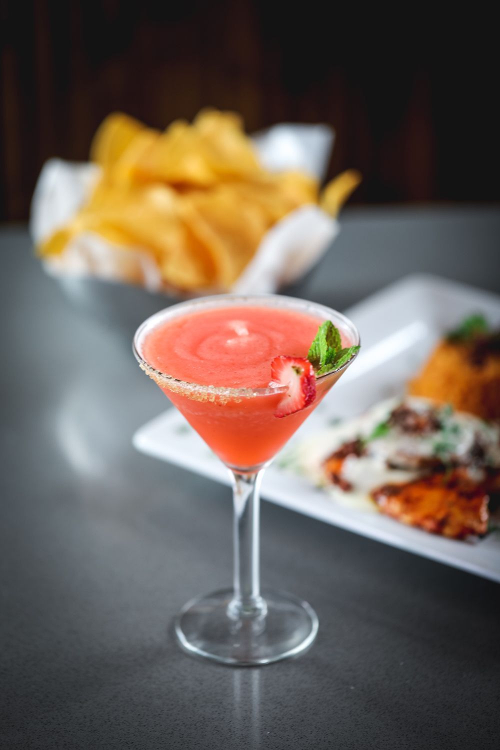 Strawberry margarita in a martini glass, garnished with a strawberry, near chips and a plate of food.