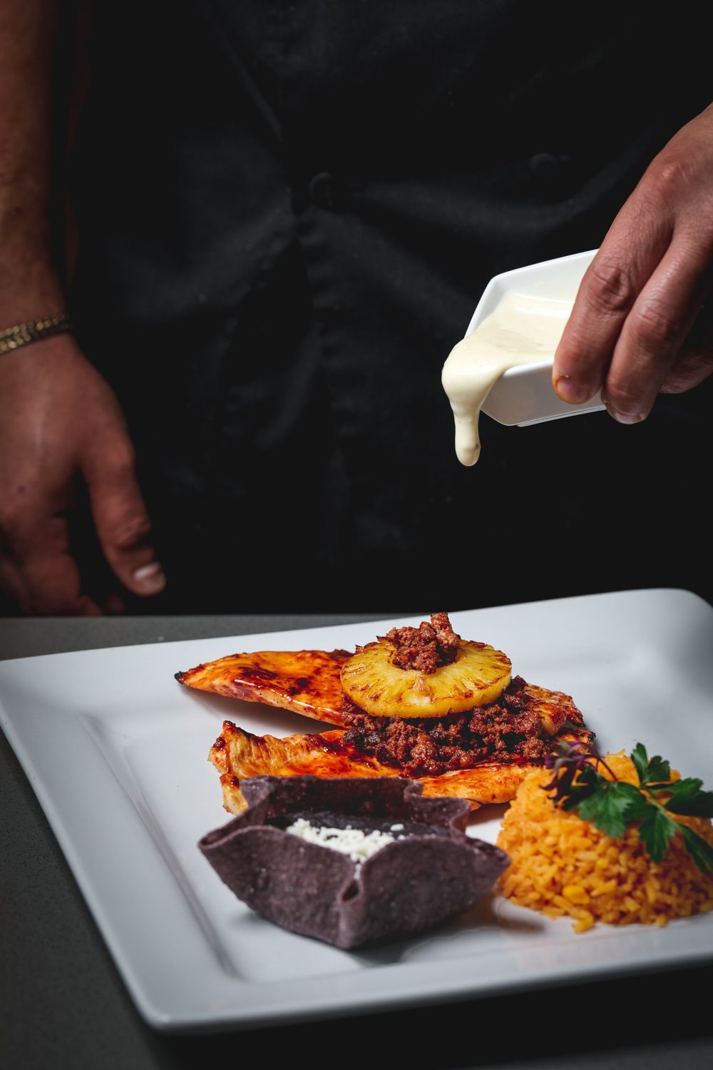 Chef drizzling white sauce over a plated dish of chicken, rice, and a blue corn tortilla cup.