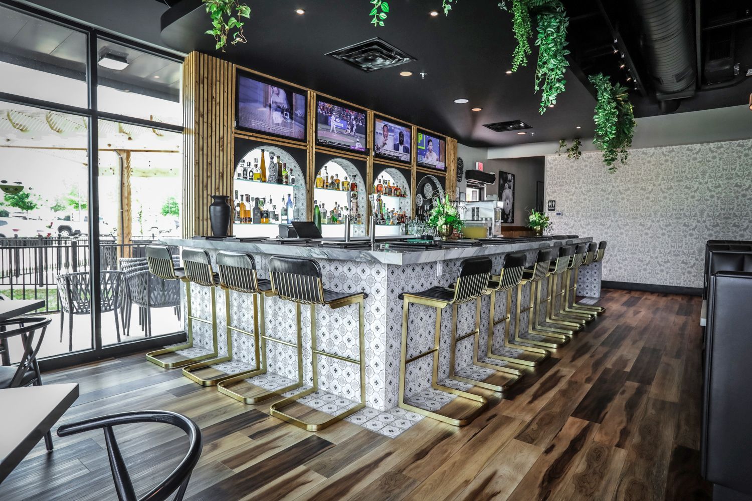 Bar with stools, TVs, and liquor displays. Wooden floor, patterned bar facade, and hanging greenery.