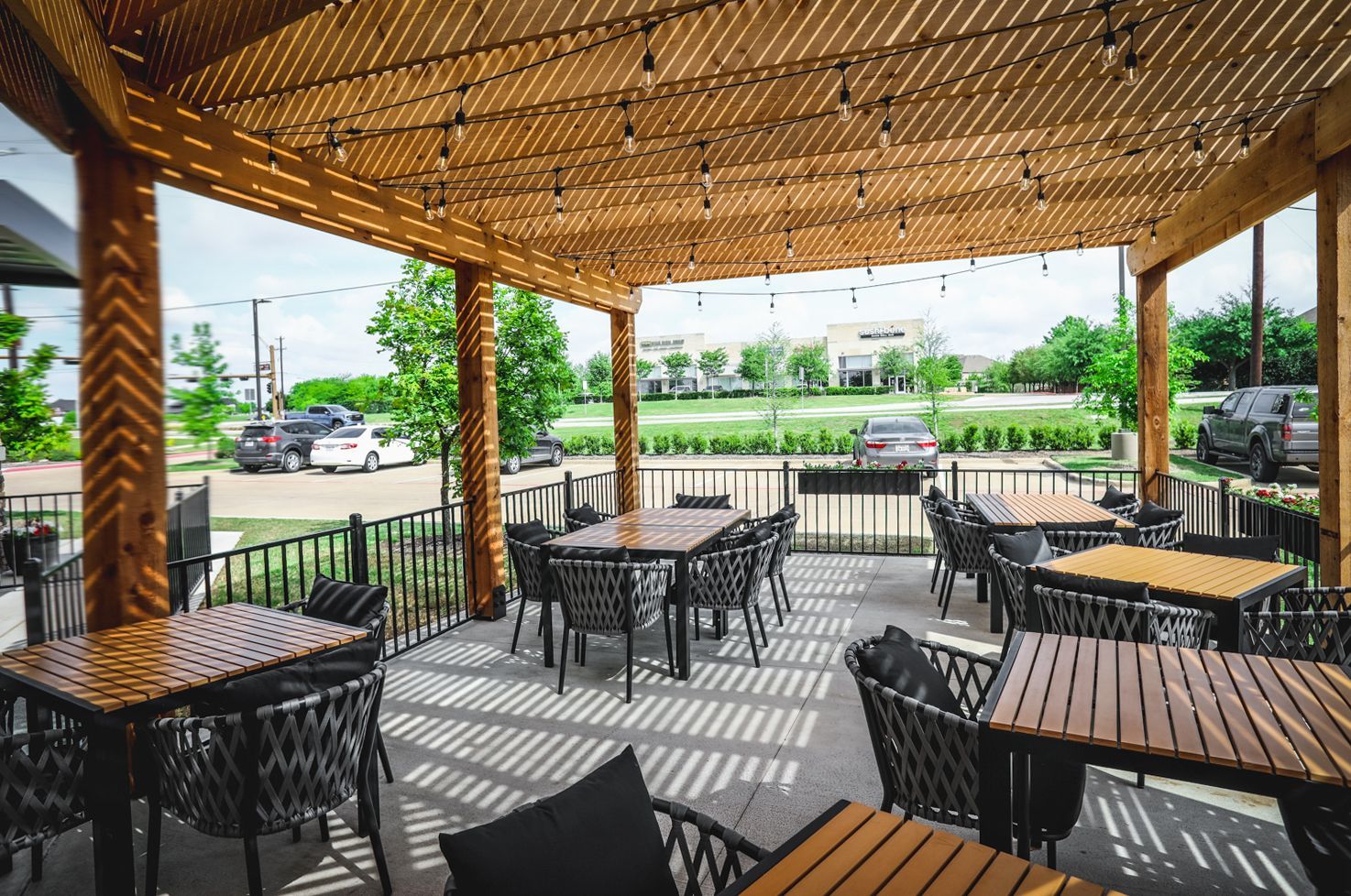 Outdoor dining patio with tables, chairs, and wooden pergola. Cars and landscaping in background.
