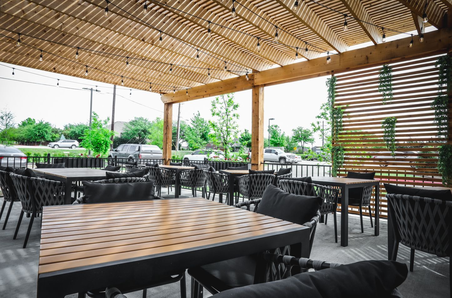 Outdoor restaurant patio with tables, chairs, and overhead wooden structure, strings of lights, overlooking a street with cars.