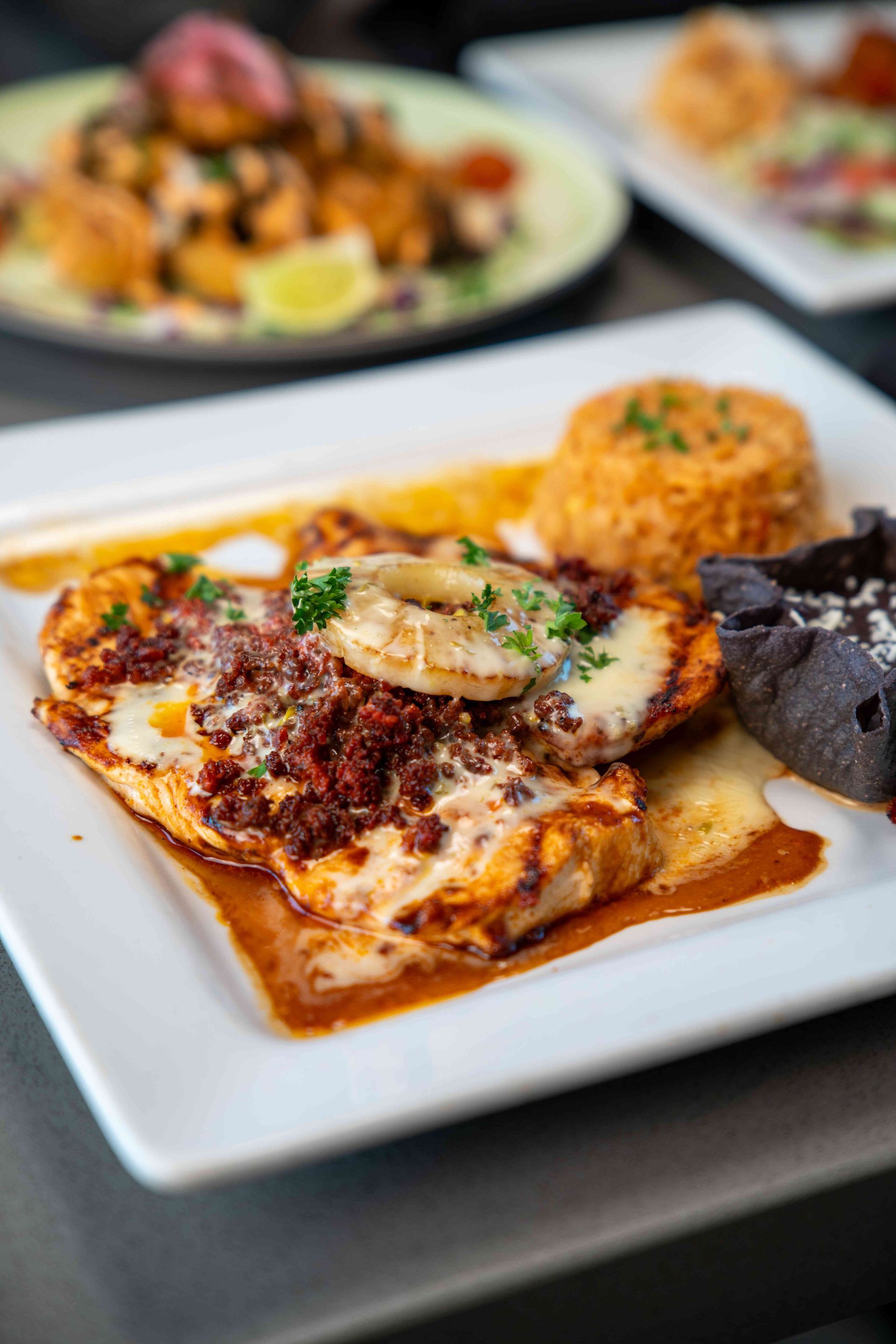 Grilled chicken breast covered in cheese and chorizo, with rice and black beans on a white plate.