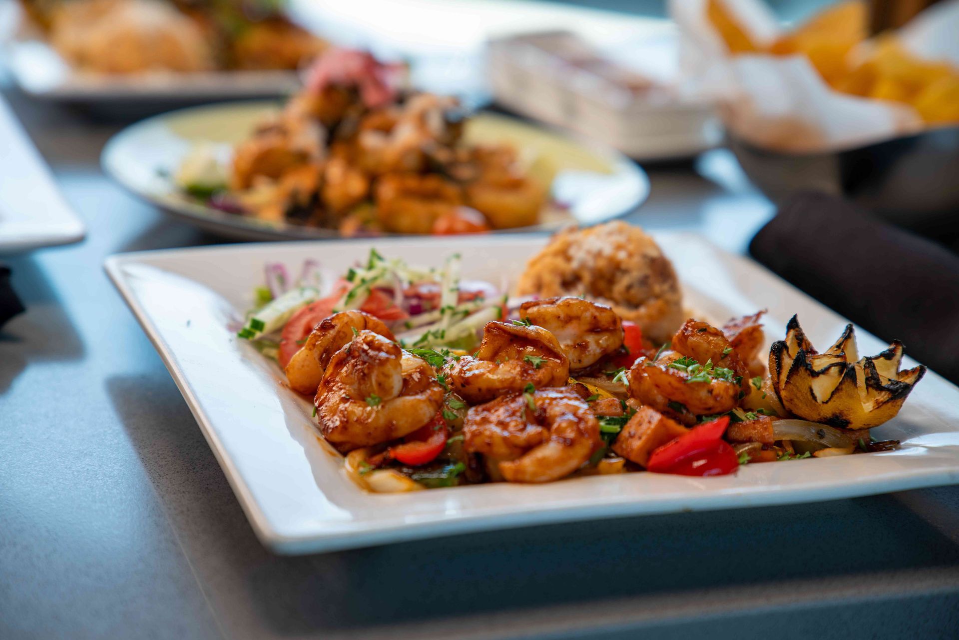 Plate of cooked shrimp with vegetables and a roll. Other plates in the background.