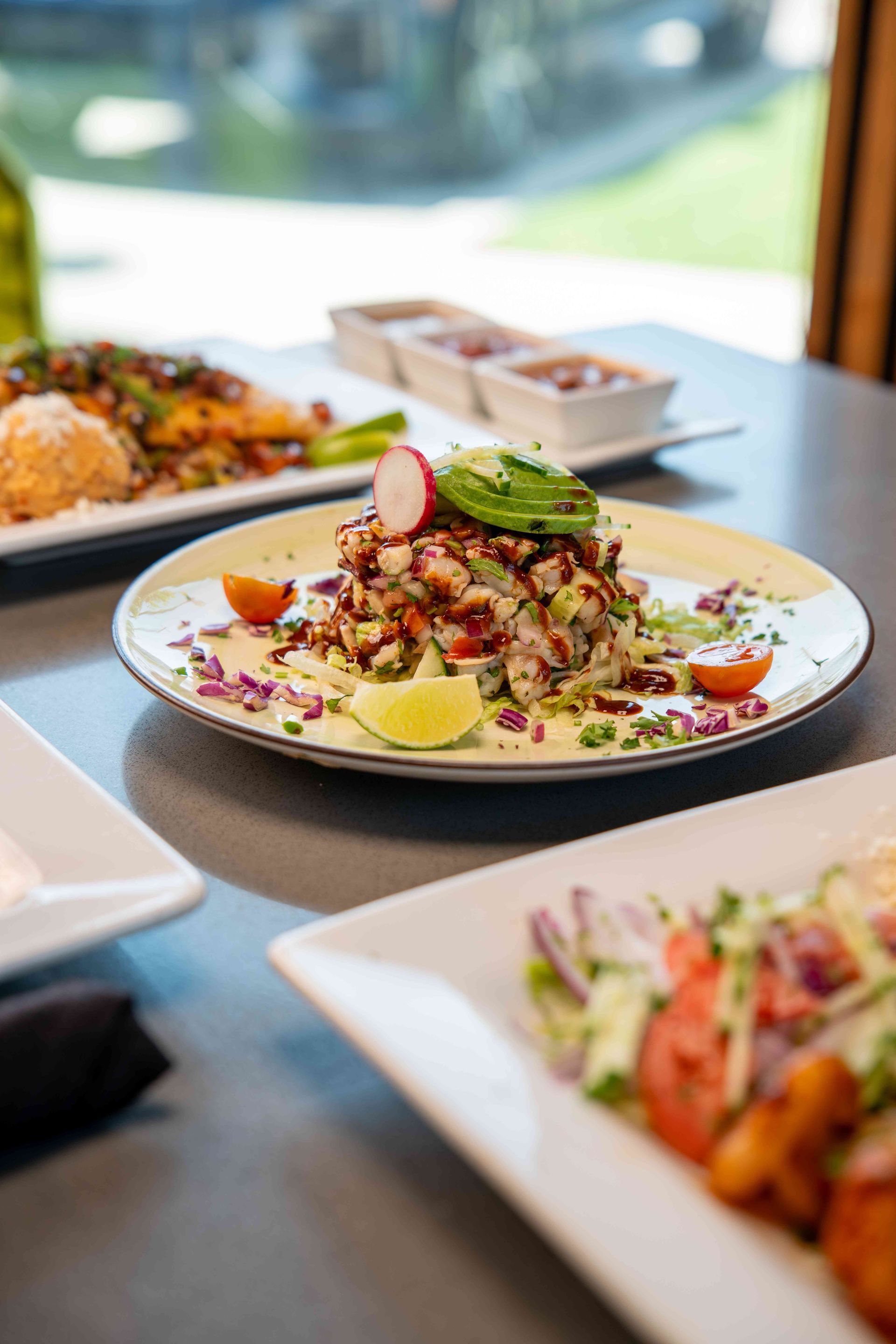 Plate of ceviche topped with avocado, radish, and lime with other dishes on a table.