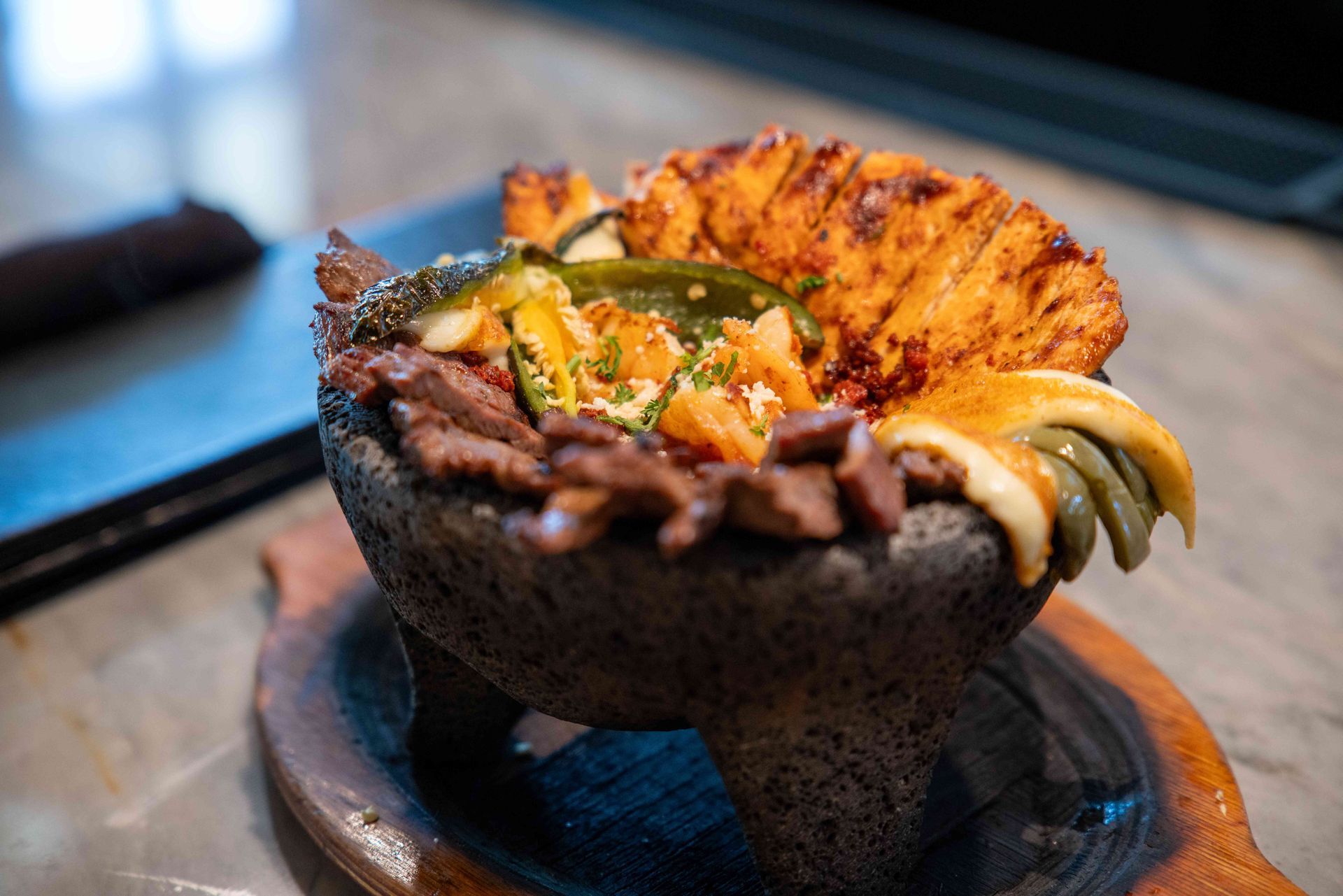 Molcajete bowl with steak, grilled vegetables, and fried cheese, on a wooden platter.