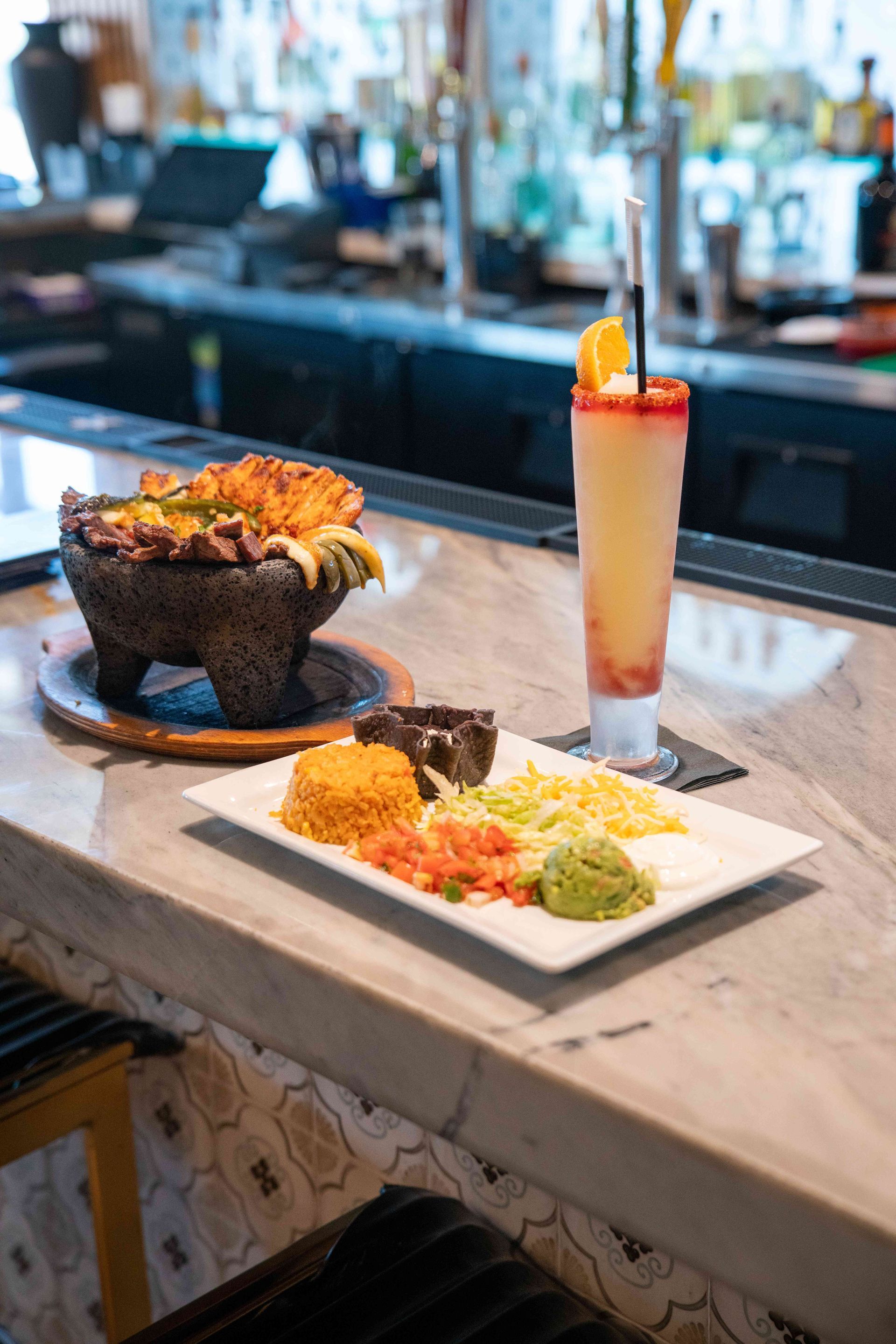 Mexican food platter with drink on a bar. Dish includes rice, salsa, guacamole, and a molcajete.