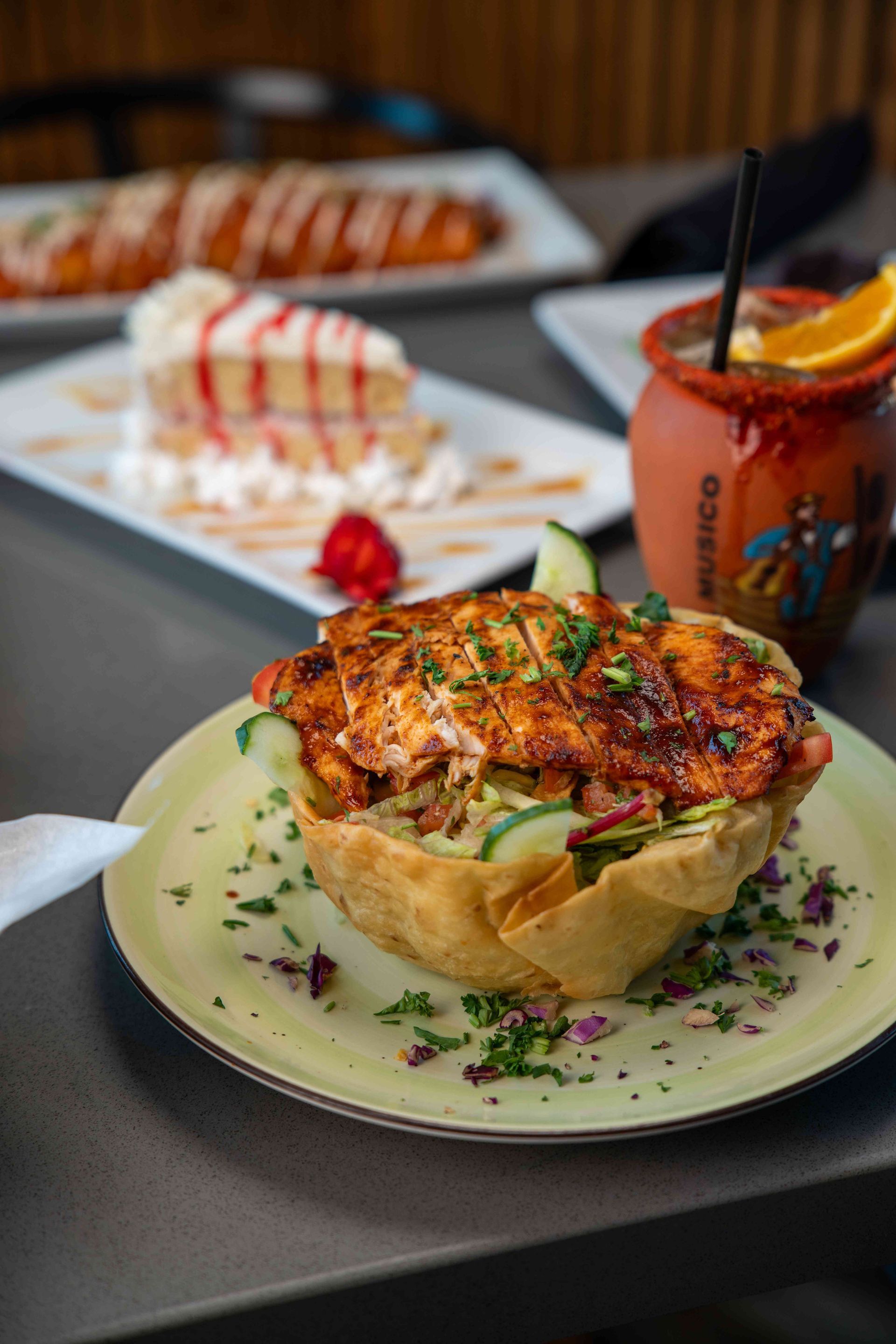 Salad in a tortilla bowl, topped with grilled chicken, with a slice of cake and a drink in the background.