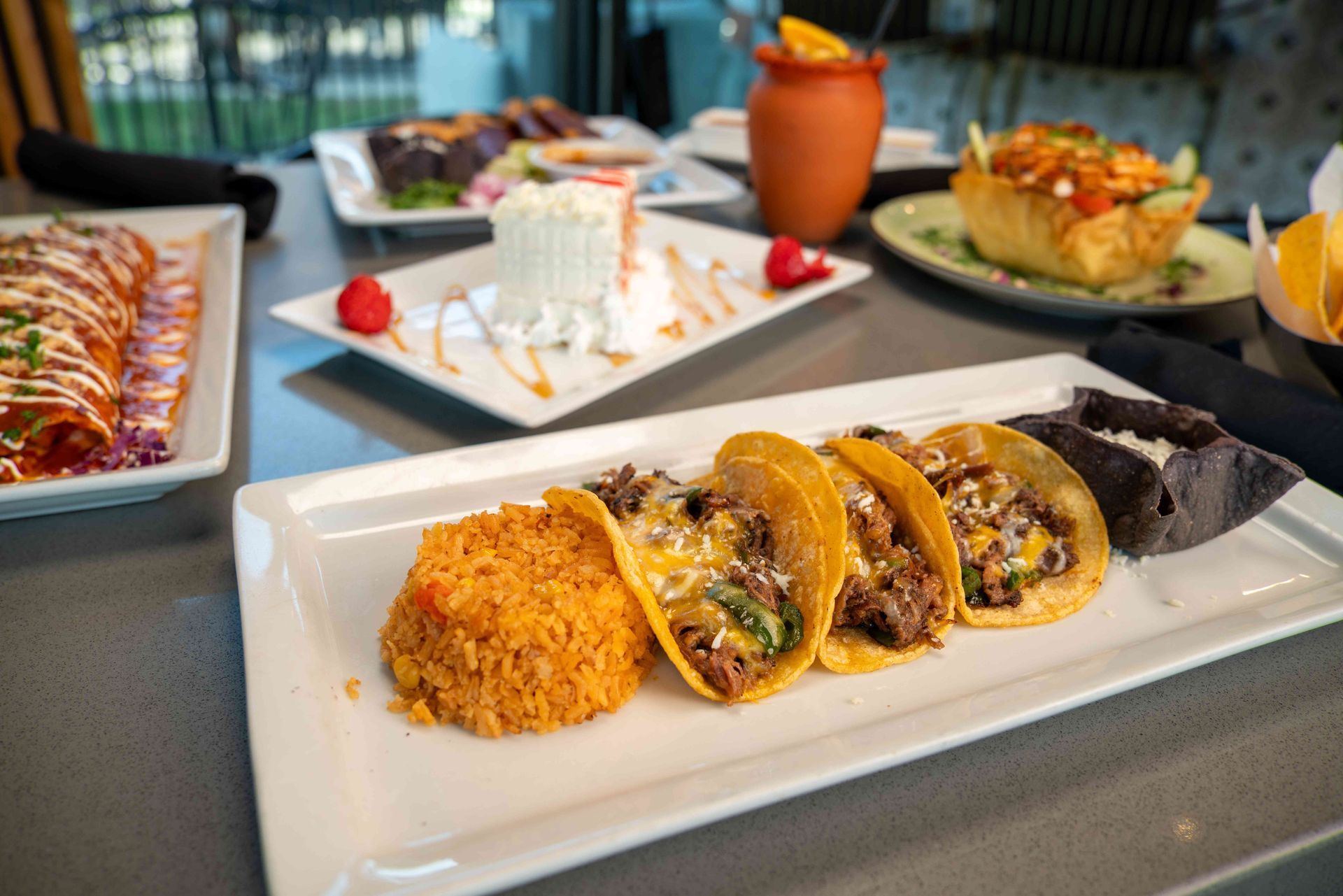 Mexican food spread: tacos, enchiladas, rice, cake, & tortilla bowl on a gray table.