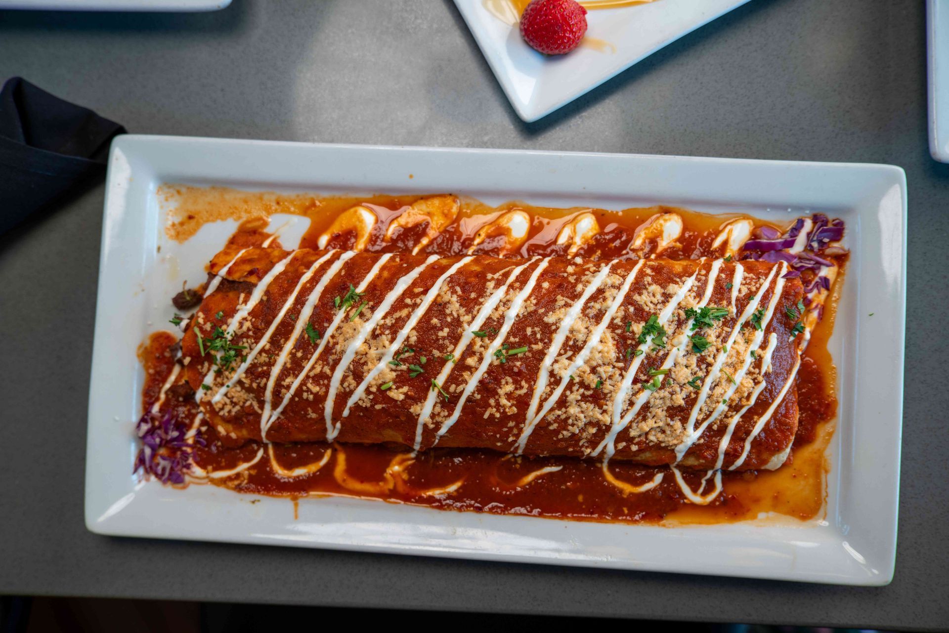 Burrito covered in red sauce and drizzle, on white platter. Garnished with shredded cabbage and herbs.