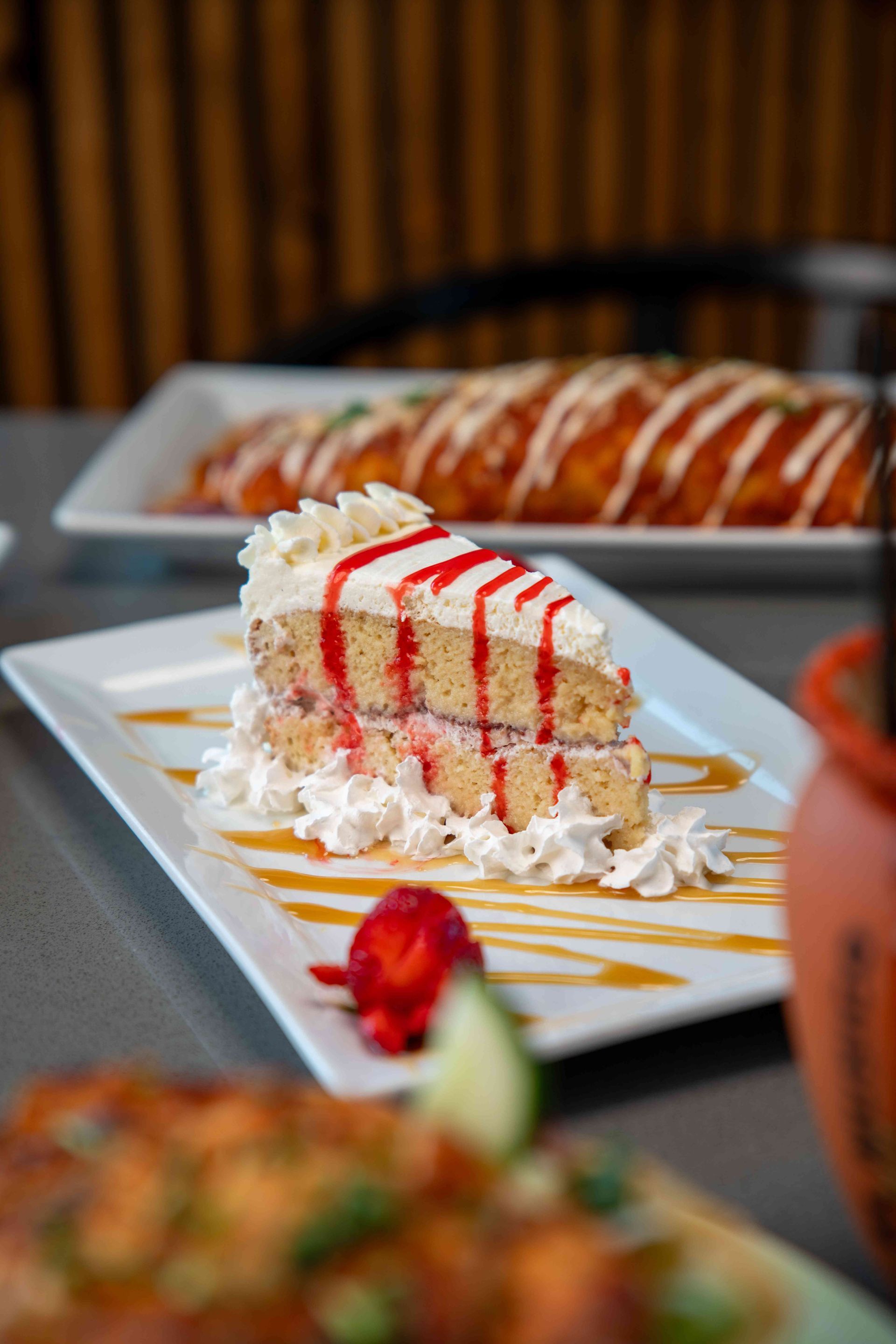 Slice of layered cake on a white plate, drizzled with red stripes and surrounded by whipped cream, next to a strawberry.