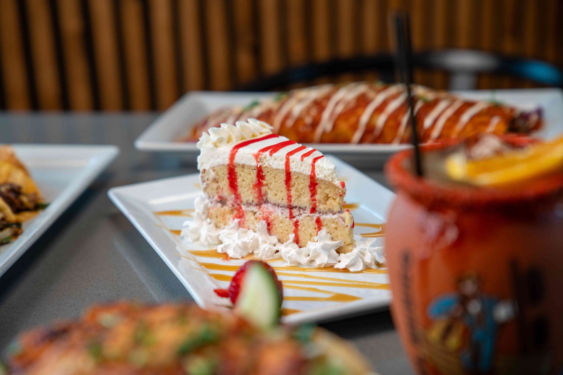 Slice of cake with whipped cream, red drizzle, and food on a table in a restaurant.
