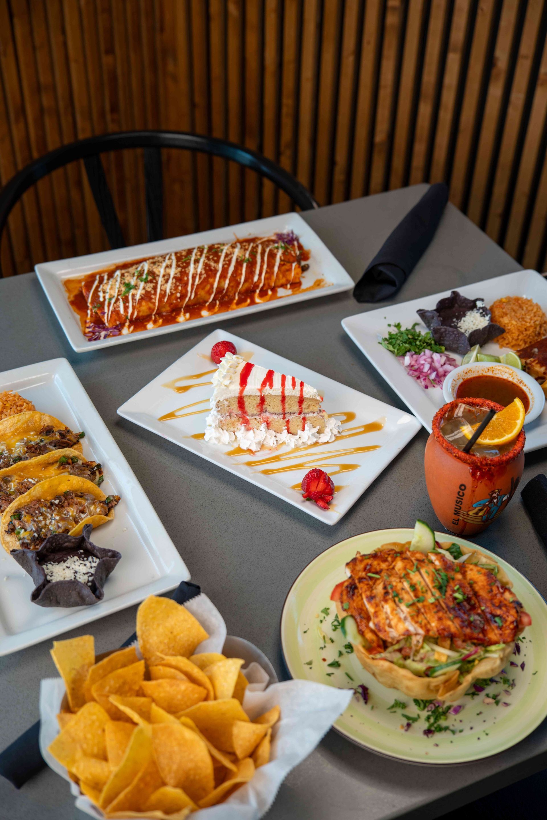 Mexican food spread on a table: enchiladas, cake, tacos, chips, and a cocktail.