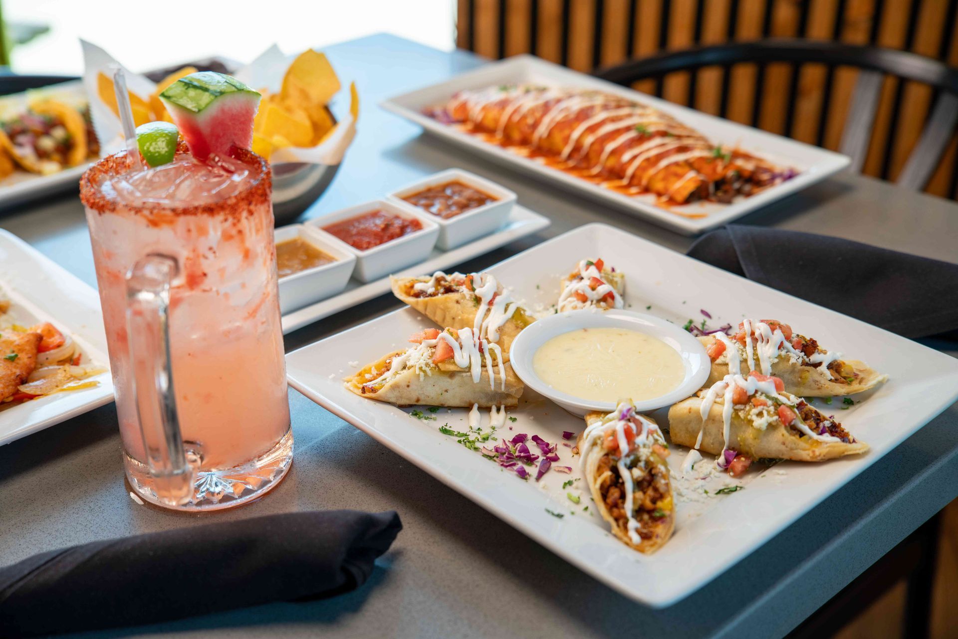 Table with Mexican food and a cocktail: tacos, enchiladas, salsa, chips, and a pink drink.