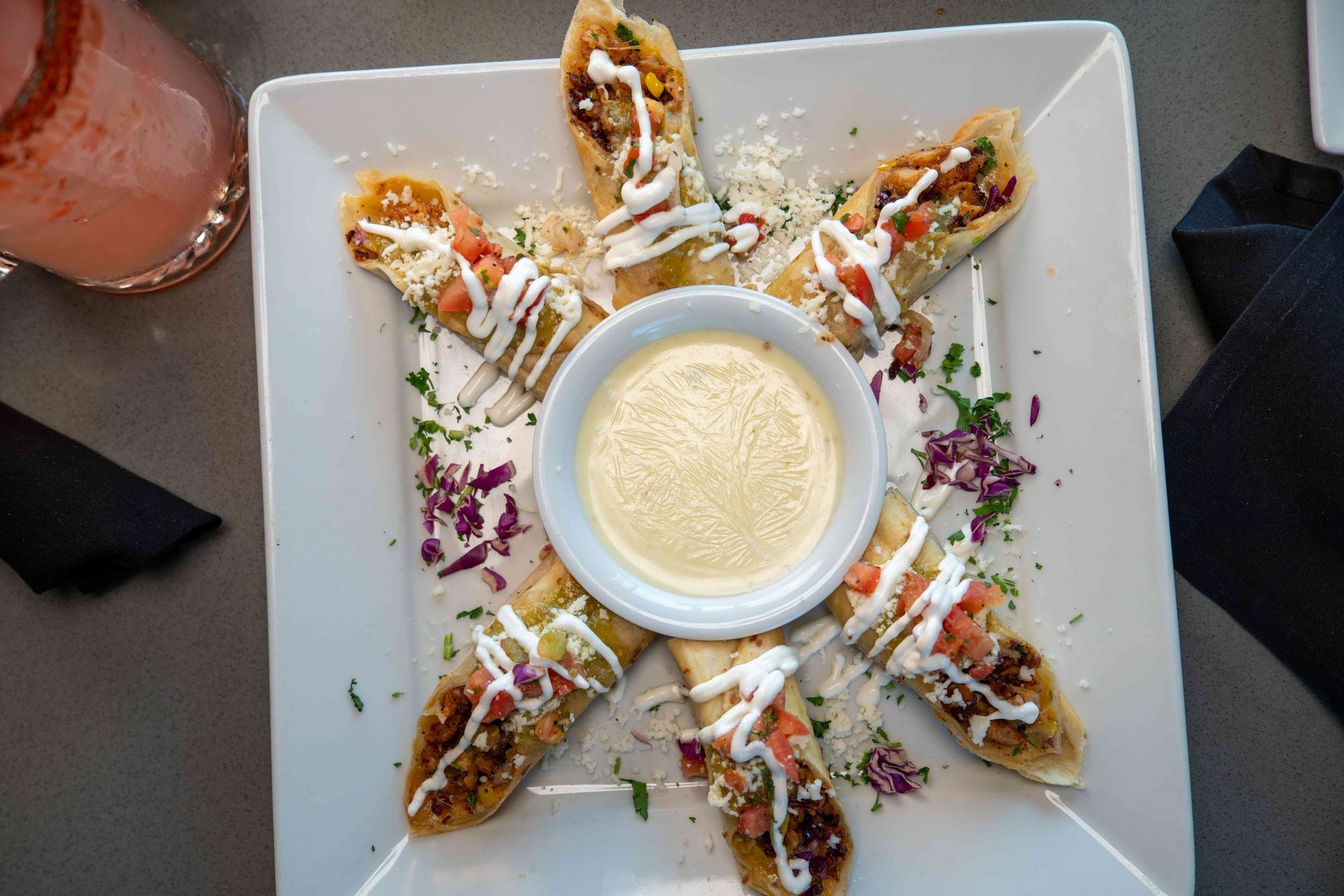 Egg rolls arranged on a square plate with dipping sauce in the center. Garnished with toppings and served with a drink.
