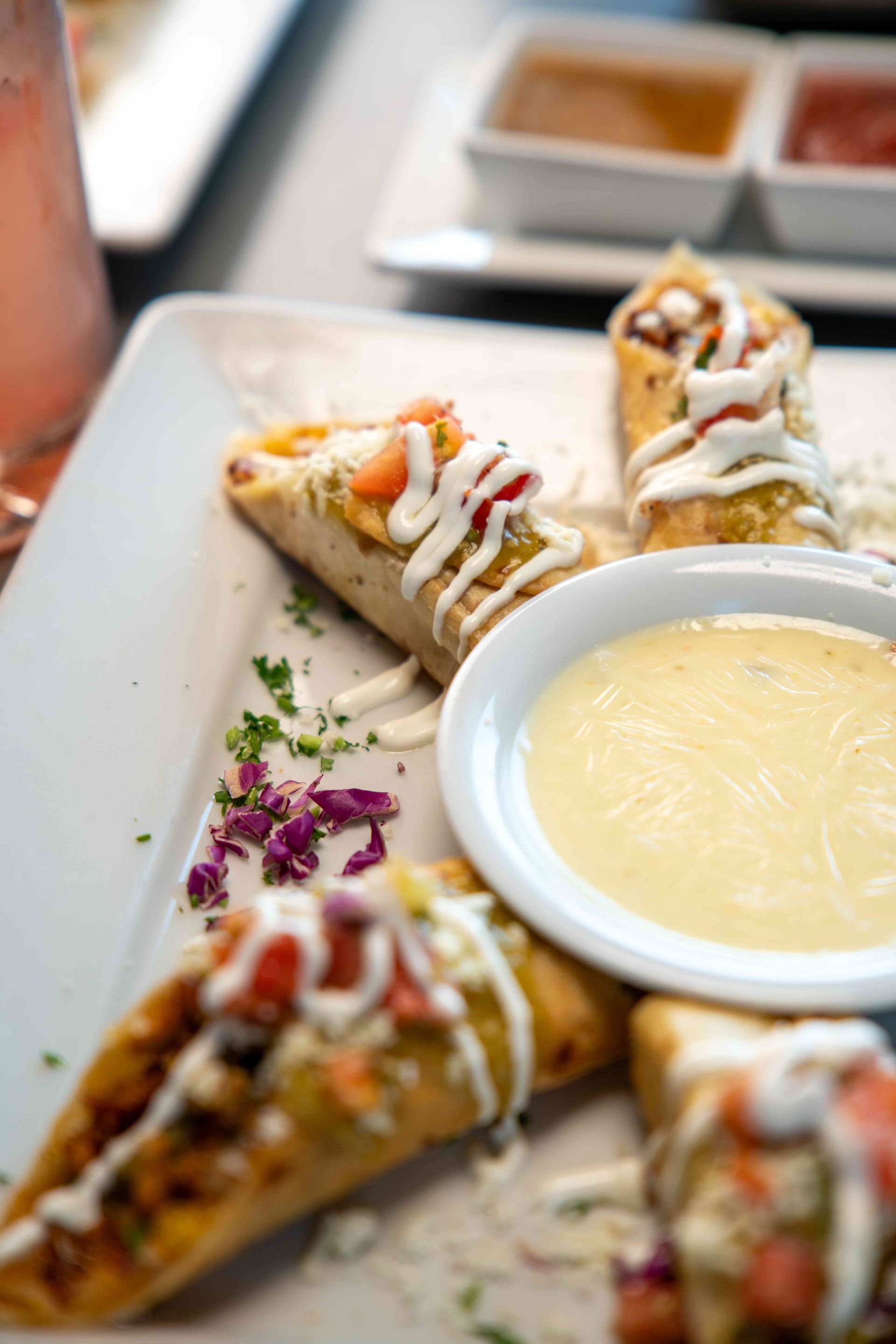 Close-up of a white plate with rolled appetizers drizzled with white sauce, salsa, and a small bowl of yellow dipping sauce.