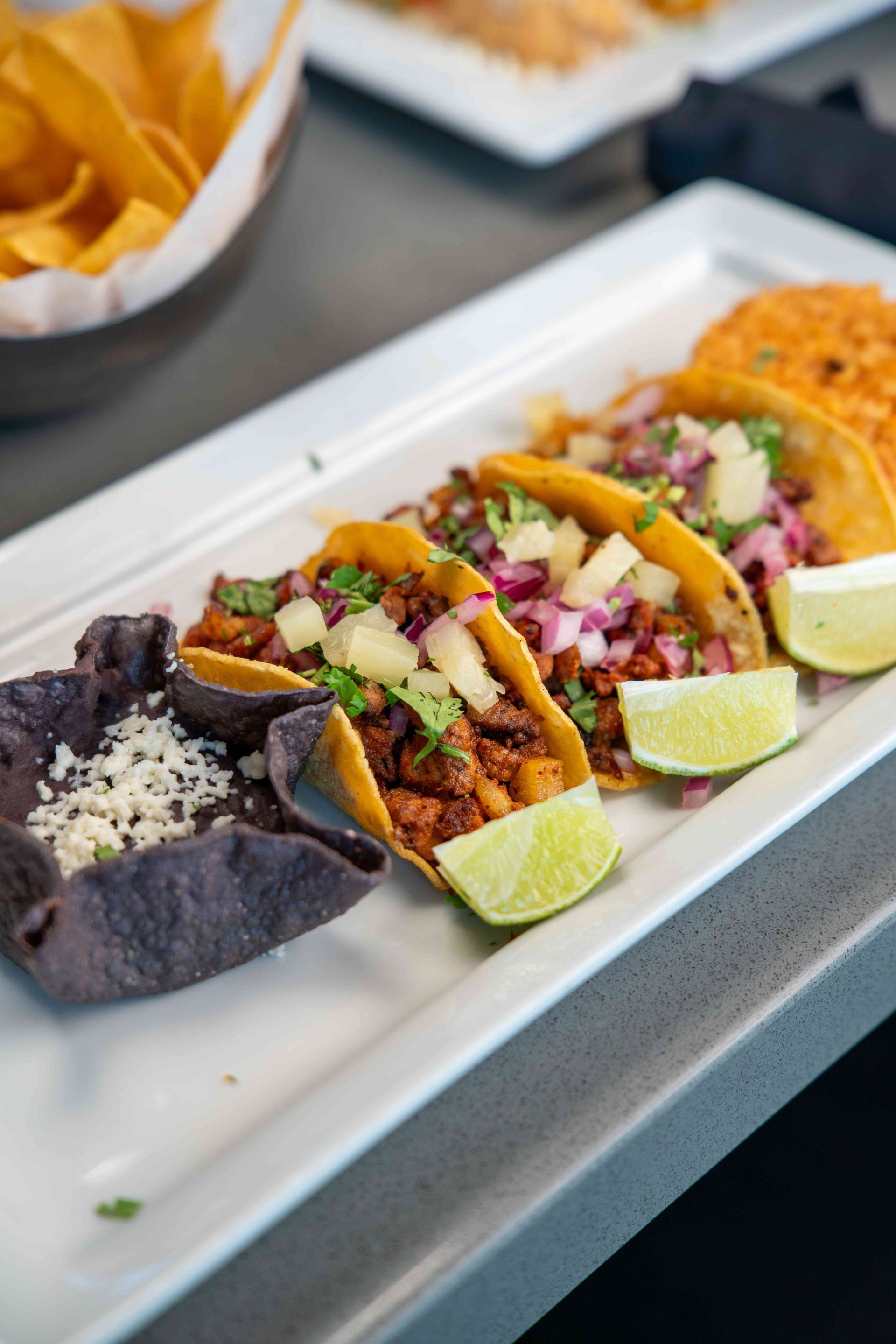Tacos with pineapple salsa, rice, black beans, tortilla chips, and lime wedges on a white platter.