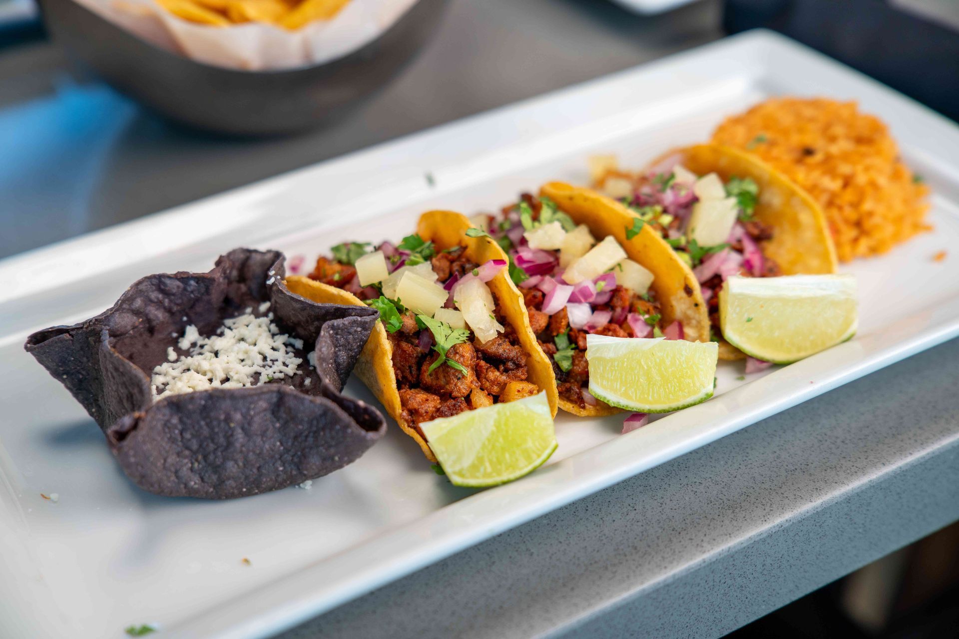 Tacos on a white plate with rice and a blue corn tortilla bowl. Includes lime wedges and pineapple.