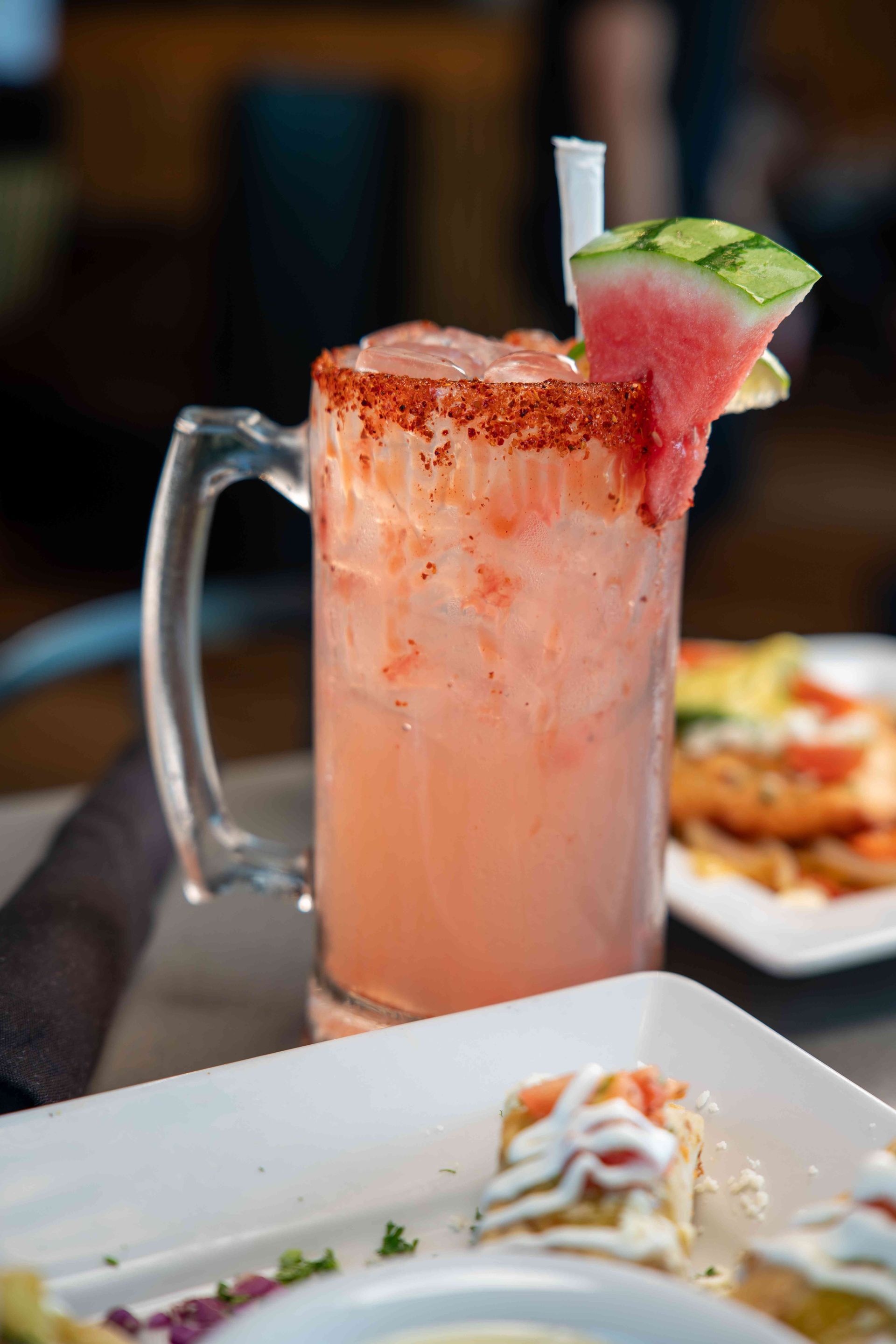 Pink cocktail in a mug rimmed with red spice, watermelon garnish, on a table with food.