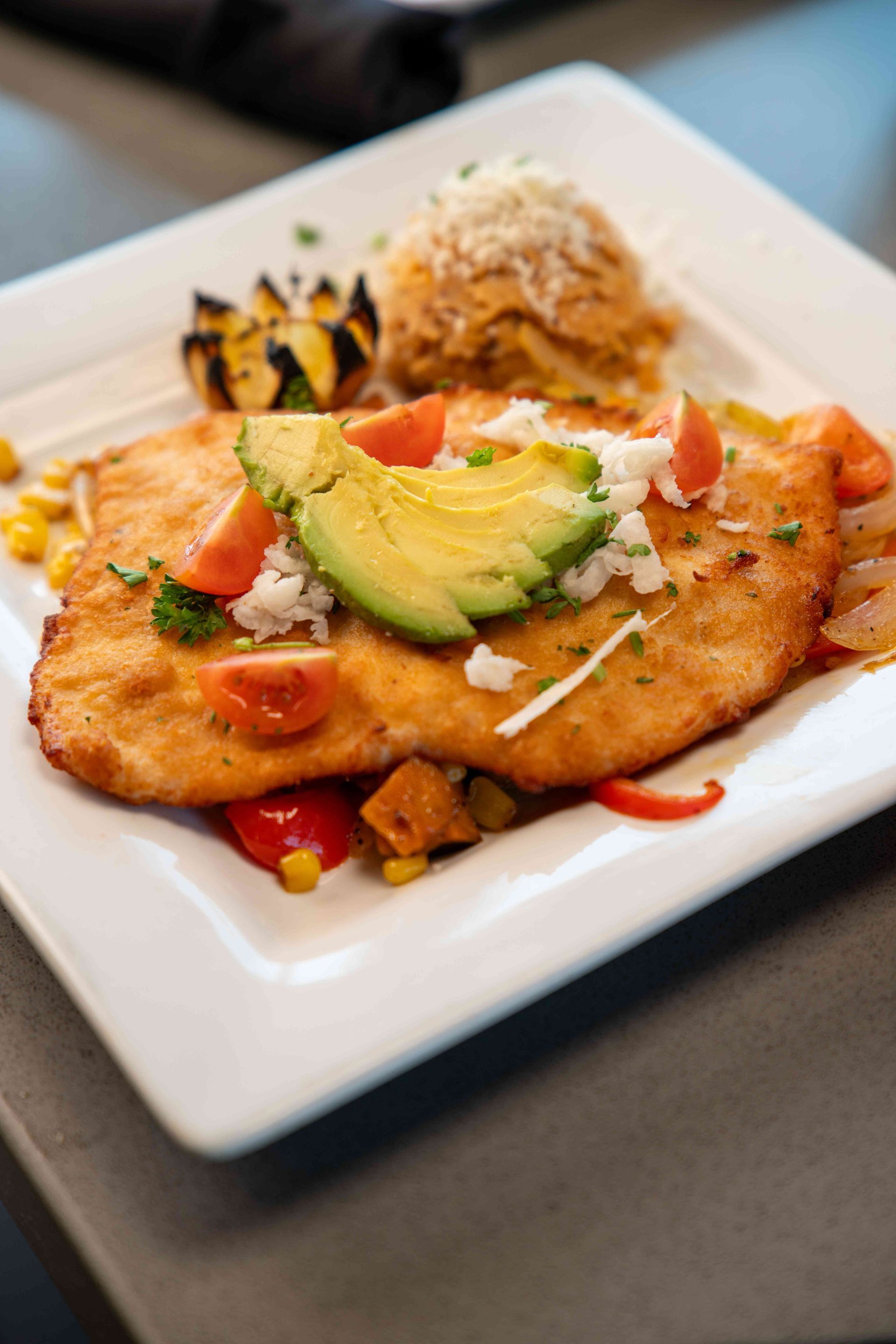 Plate of fried fish with avocado, tomatoes, rice, and grilled vegetables.