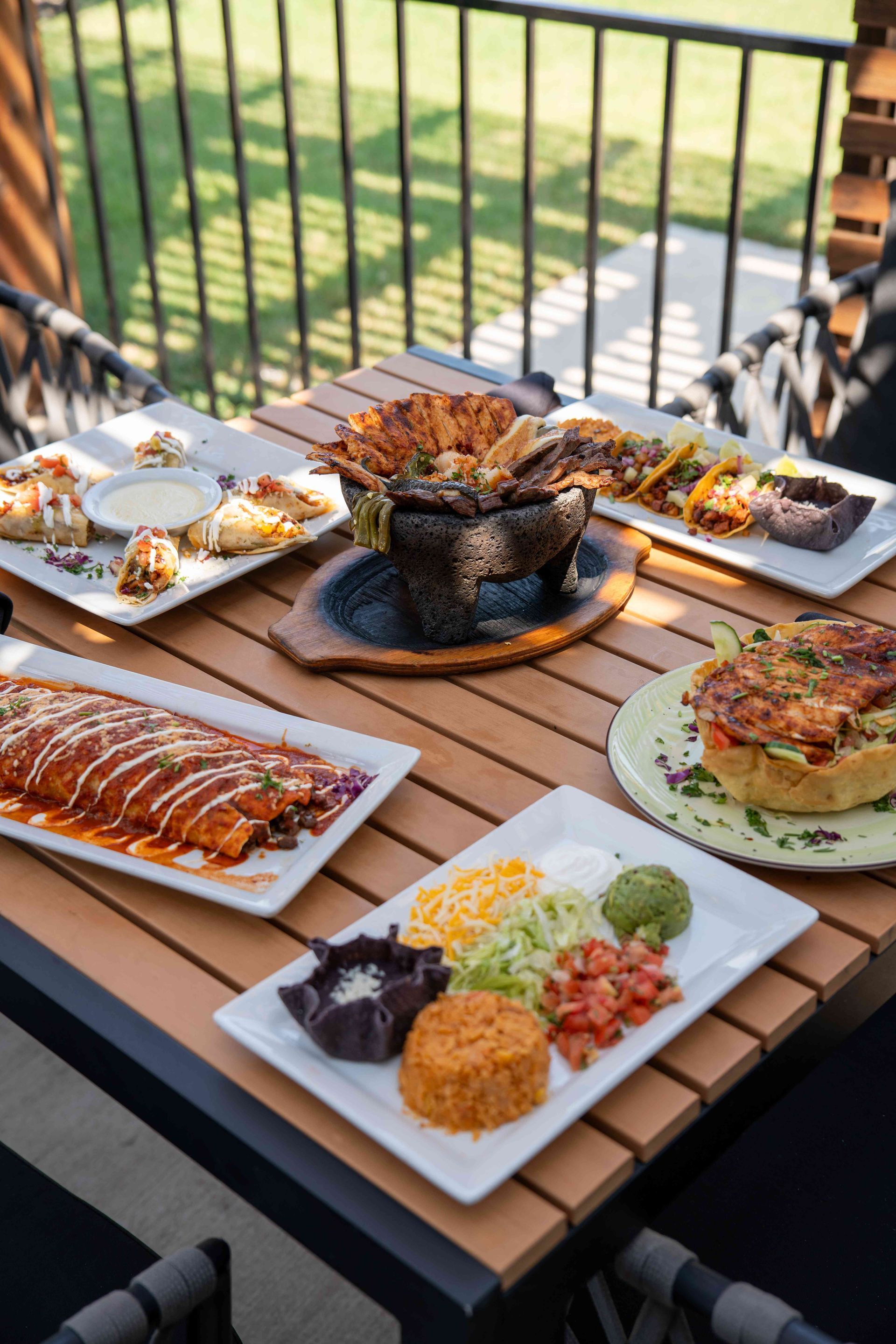 Various Mexican dishes on a wooden table outdoors: enchiladas, tacos, rice, guacamole, and more.
