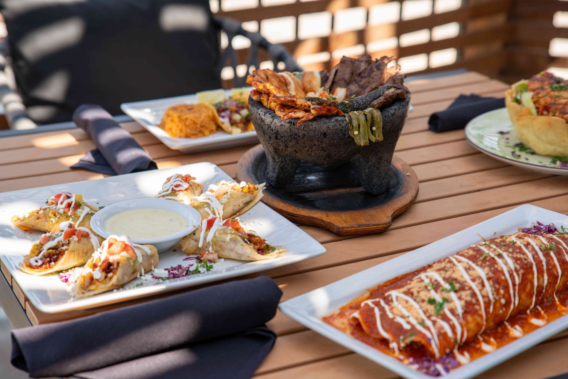 Mexican food spread on a wooden table, including tacos, burritos, and a molcajete with meat.