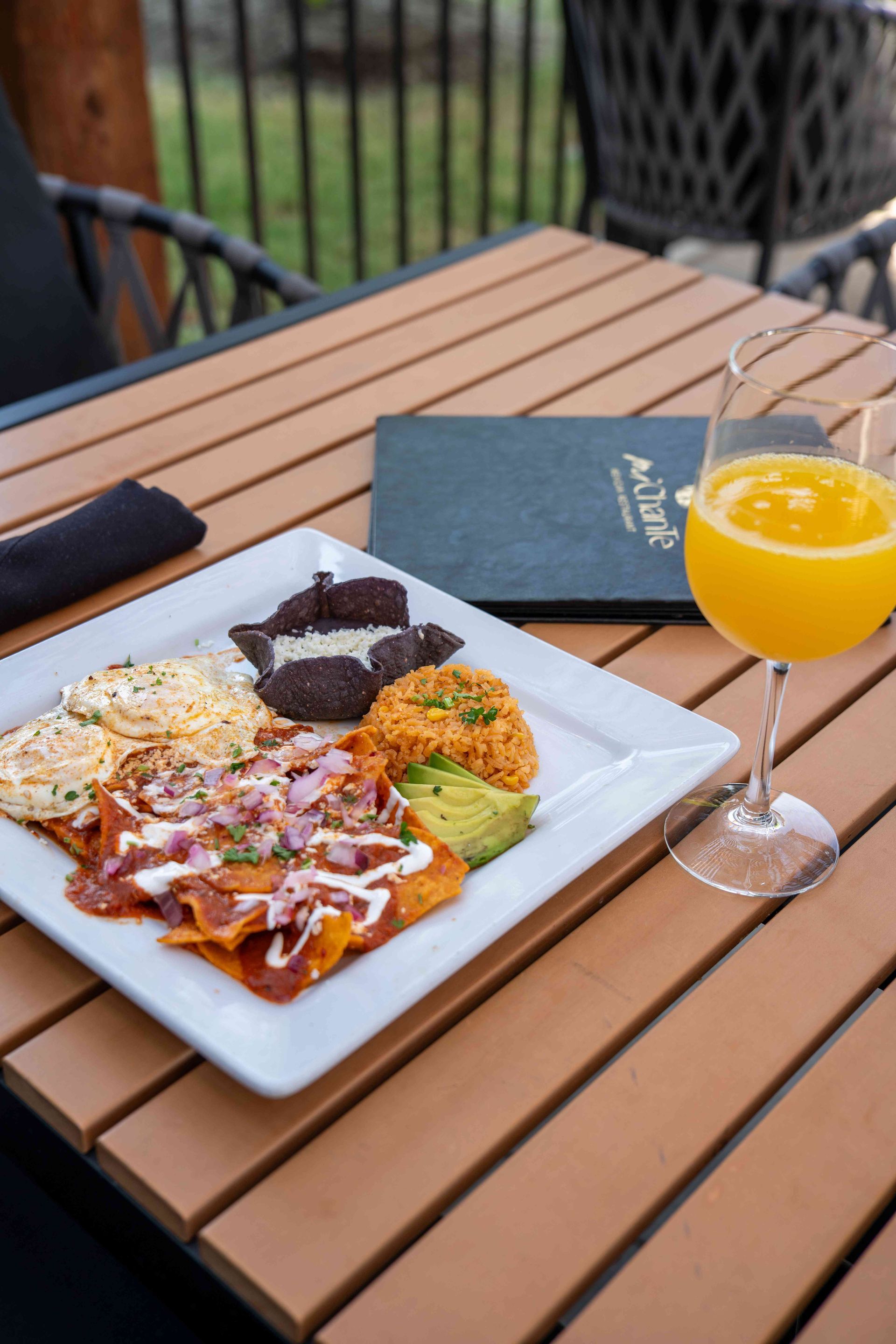 Plate of food and mimosa on a wooden table outdoors. Dish includes chilaquiles, black beans, avocado, and rice.