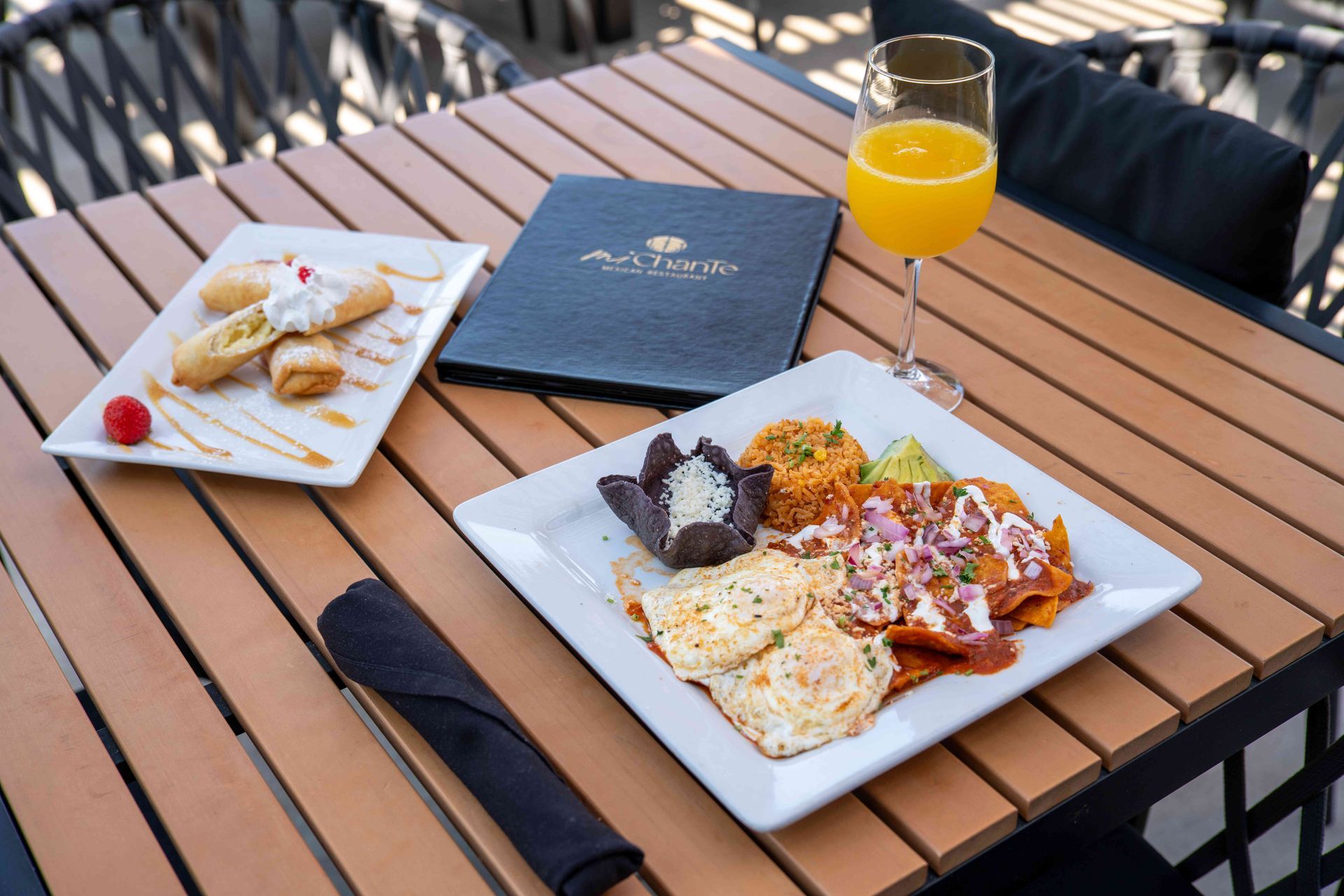 Table with food: enchiladas, dessert, mimosa, and menu. Outdoor setting.