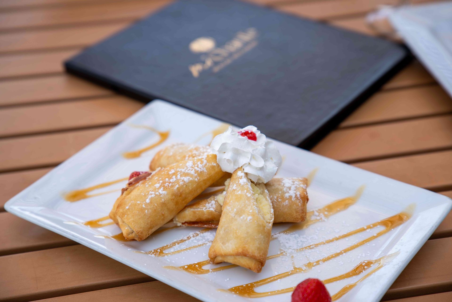Fried dessert rolls on a white plate, drizzled with caramel sauce, whipped cream, and powdered sugar, menu in the background.