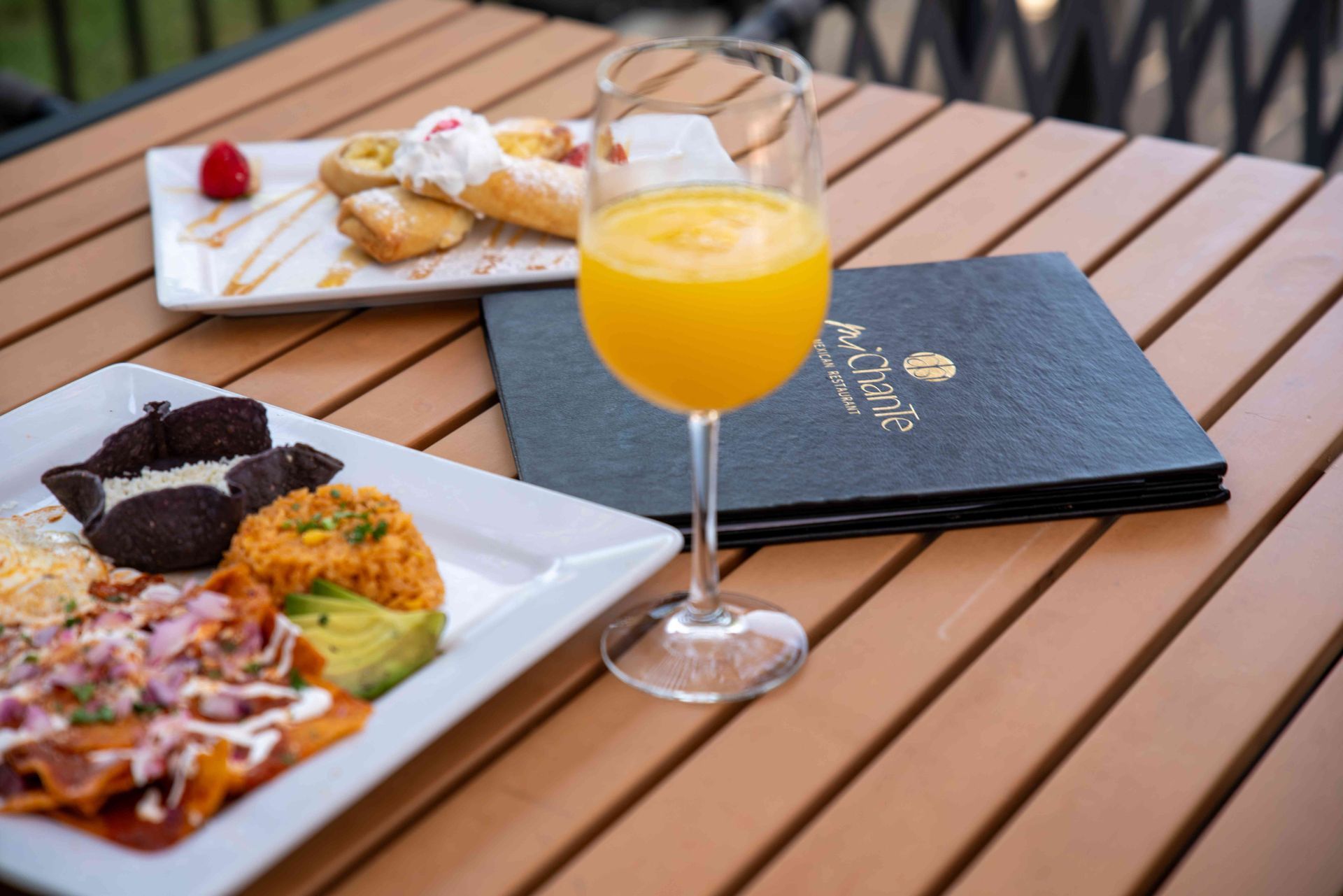 Brunch table with mimosa, menu, and plates of food: sweet pastry, and savory Mexican dishes.