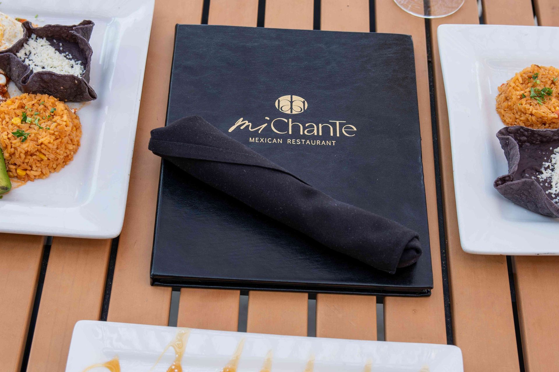 Menu and food plates on a wooden table. The menu is black with gold lettering for 