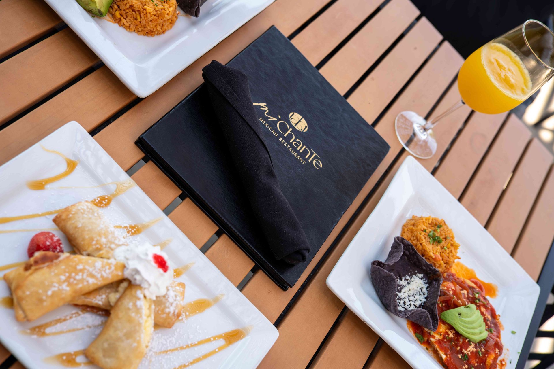 Table of food at a restaurant: Fried pastries, entrees with sauces, mimosa, and a menu.